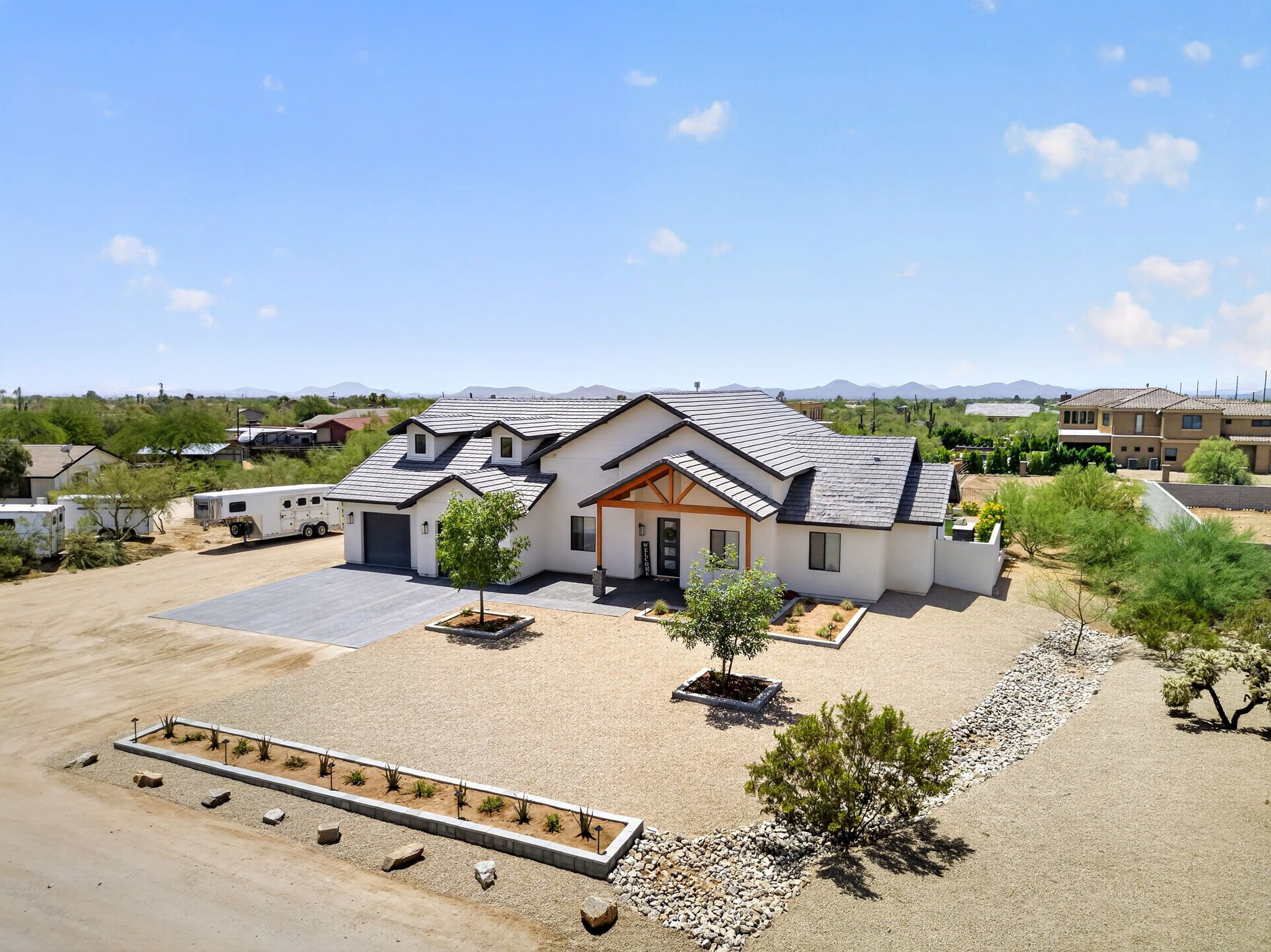 View of a new modern house with a landscaped yard, small trees, gravel paths, and distant mountains under a partly cloudy sky.