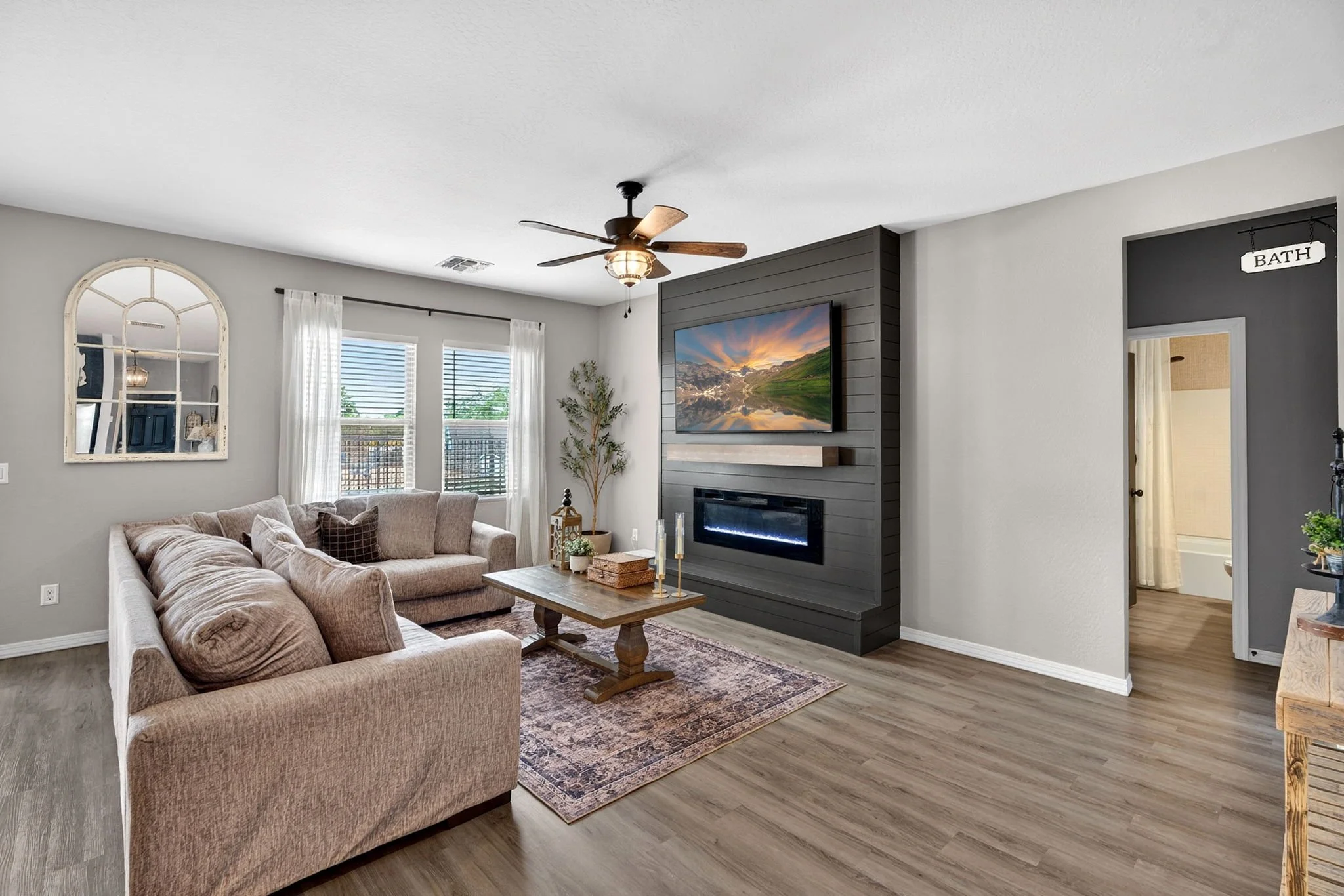 Living room with beige sectional sofa, coffee table, wall-mounted television, fireplace, window with sheer curtains, ceiling fan, and a door to a bathroom.