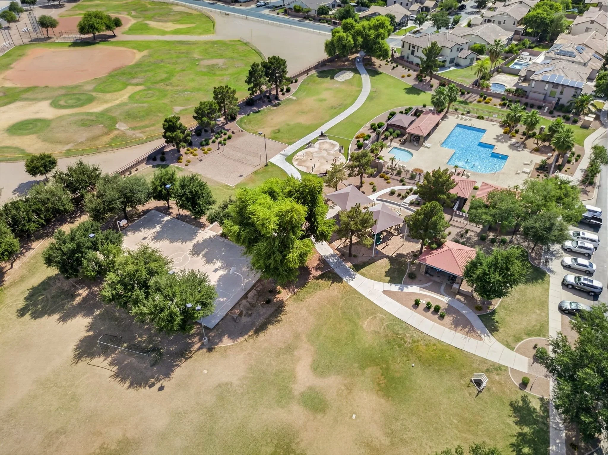 Aerial view of a neighborhood park area with open grassy fields, walking paths, a playground, and a swimming pool area surrounded by residential houses.