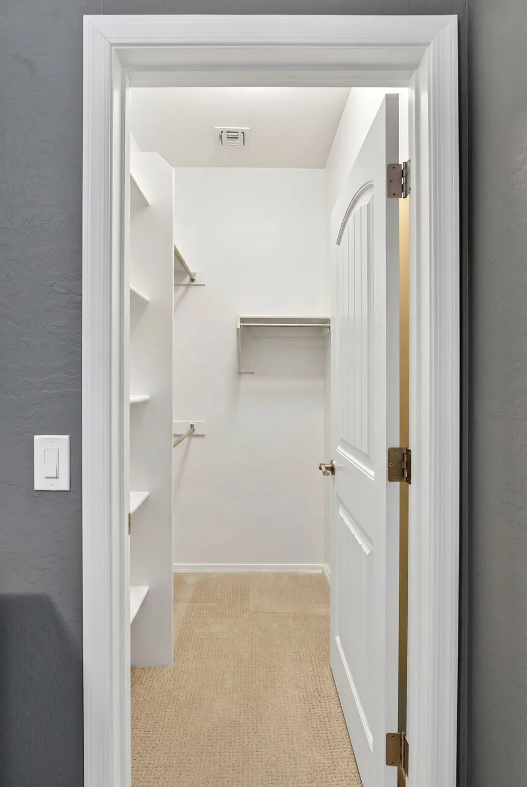 Open door revealing a white walk-in closet with shelves and hanging rods, carpeted floor, and a vent on the ceiling.