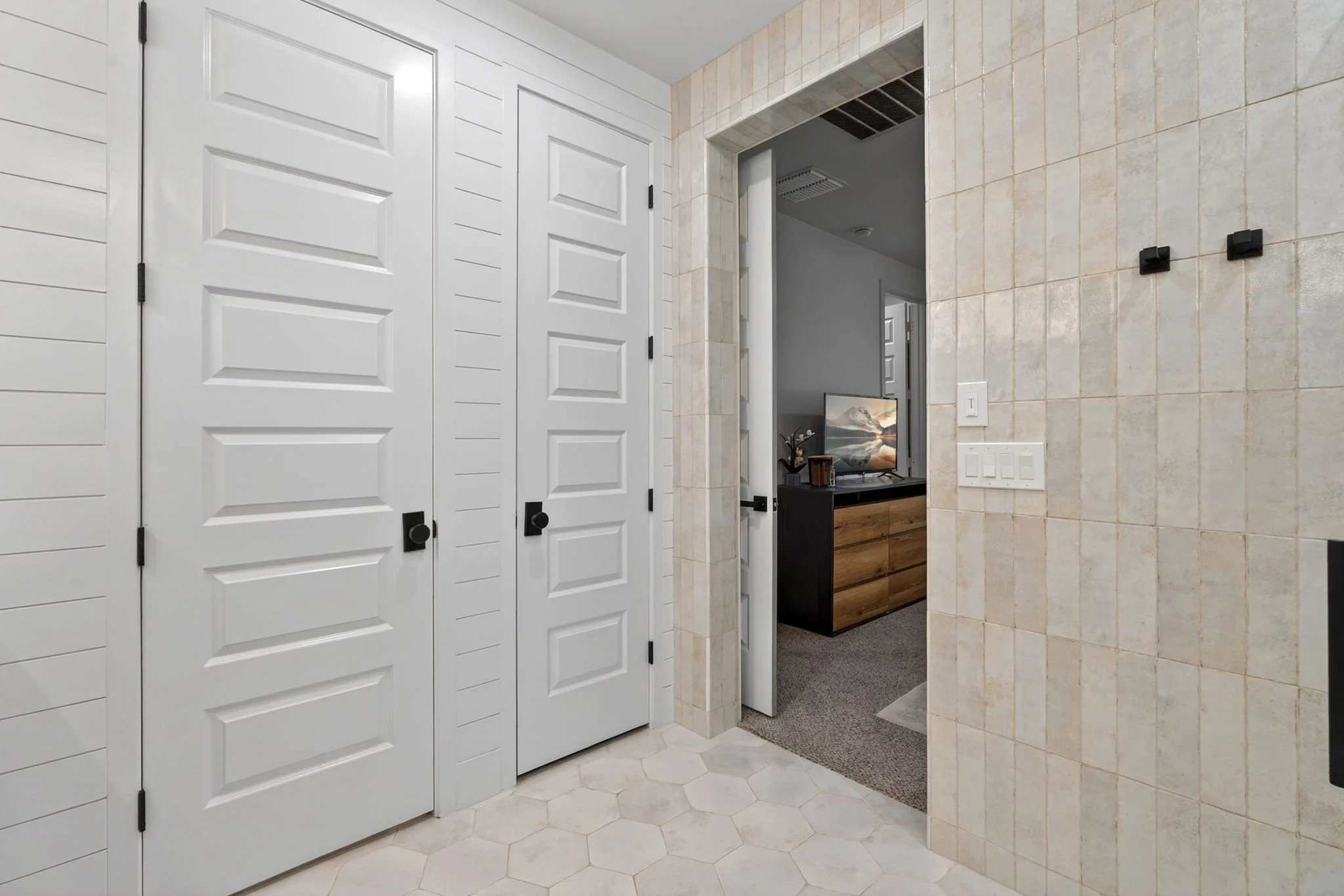 Interior view of a room with white walls and louvered closet doors, tiled walls, a small TV on a wooden cabinet, and a hallway visible through an open door.