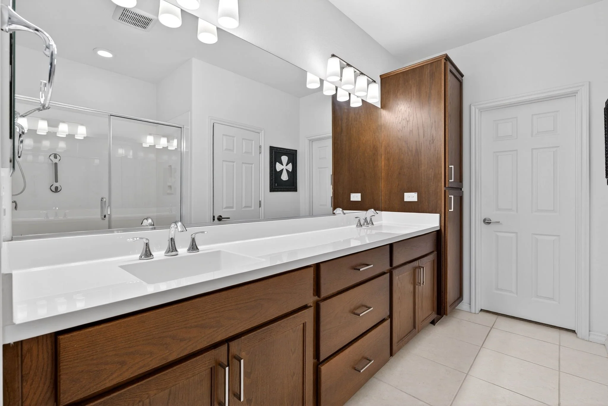Bathroom vanity with a white countertop, wooden cabinets, and a large mirror. The bathroom has a shower with glass doors and a framed artwork on the wall.