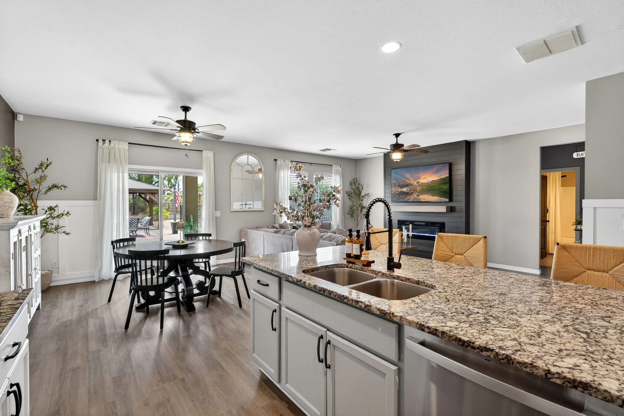Open-concept living room and kitchen with granite island, white cabinetry, and a wall-mounted flat-screen TV, decorated with plants, white curtains, and ceiling fans.