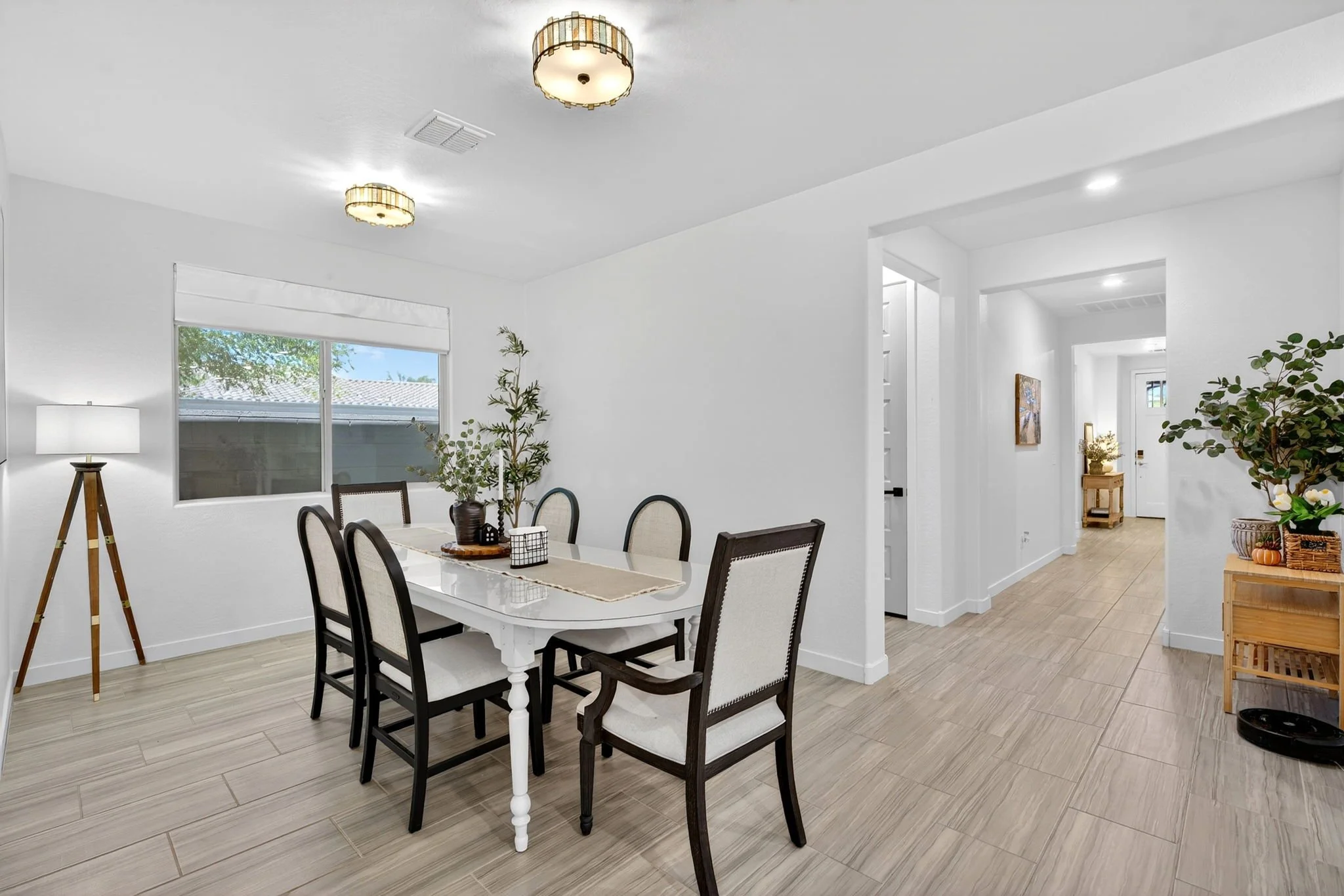 A dining room with a white table and six chairs, a window with blinds, a floor lamp, indoor plants, and hallway with artwork and more plants.
