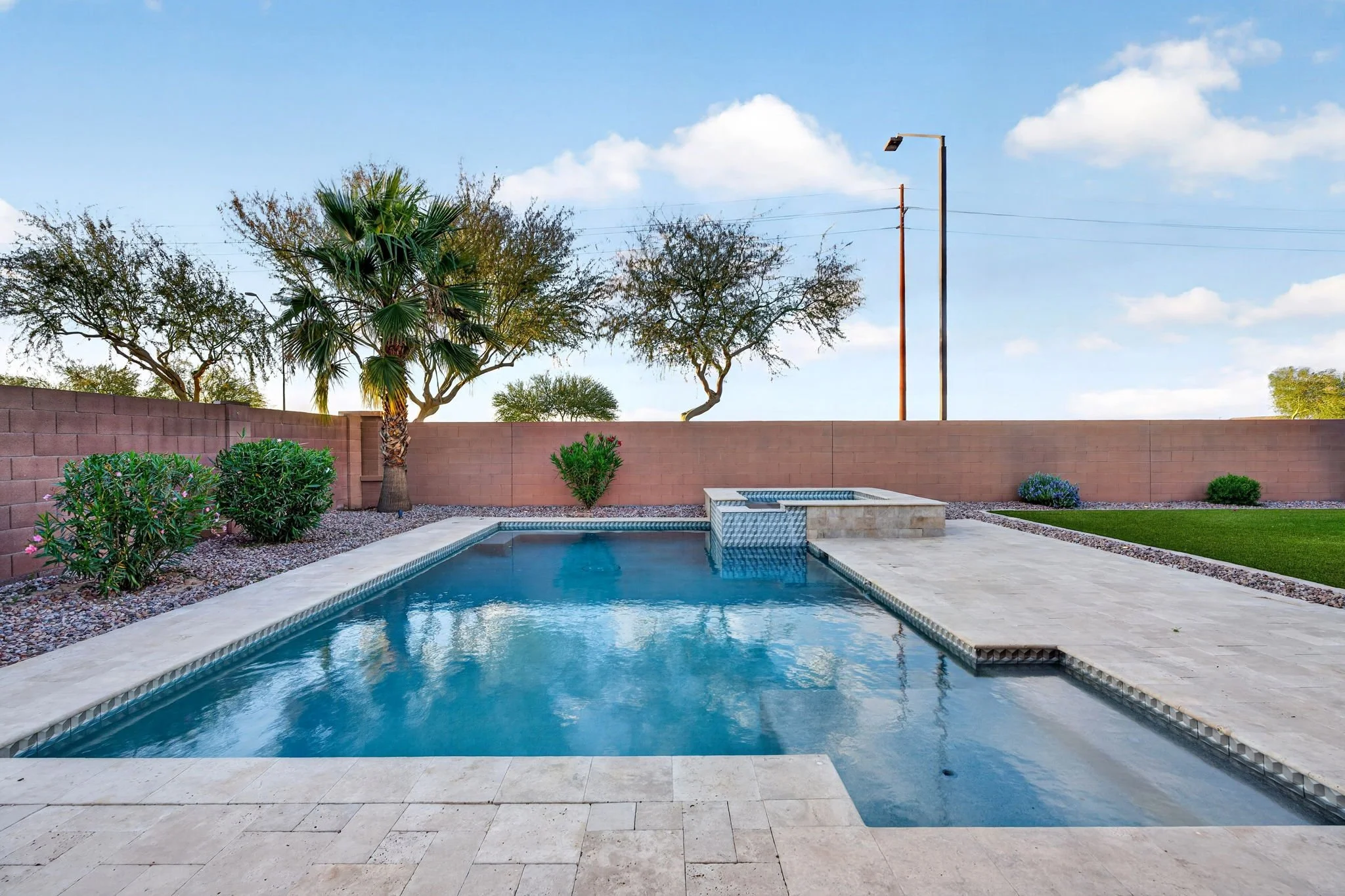 Backyard with a swimming pool, desert landscaping, a tall brick wall, palm and other desert trees, and a cloudy blue sky.