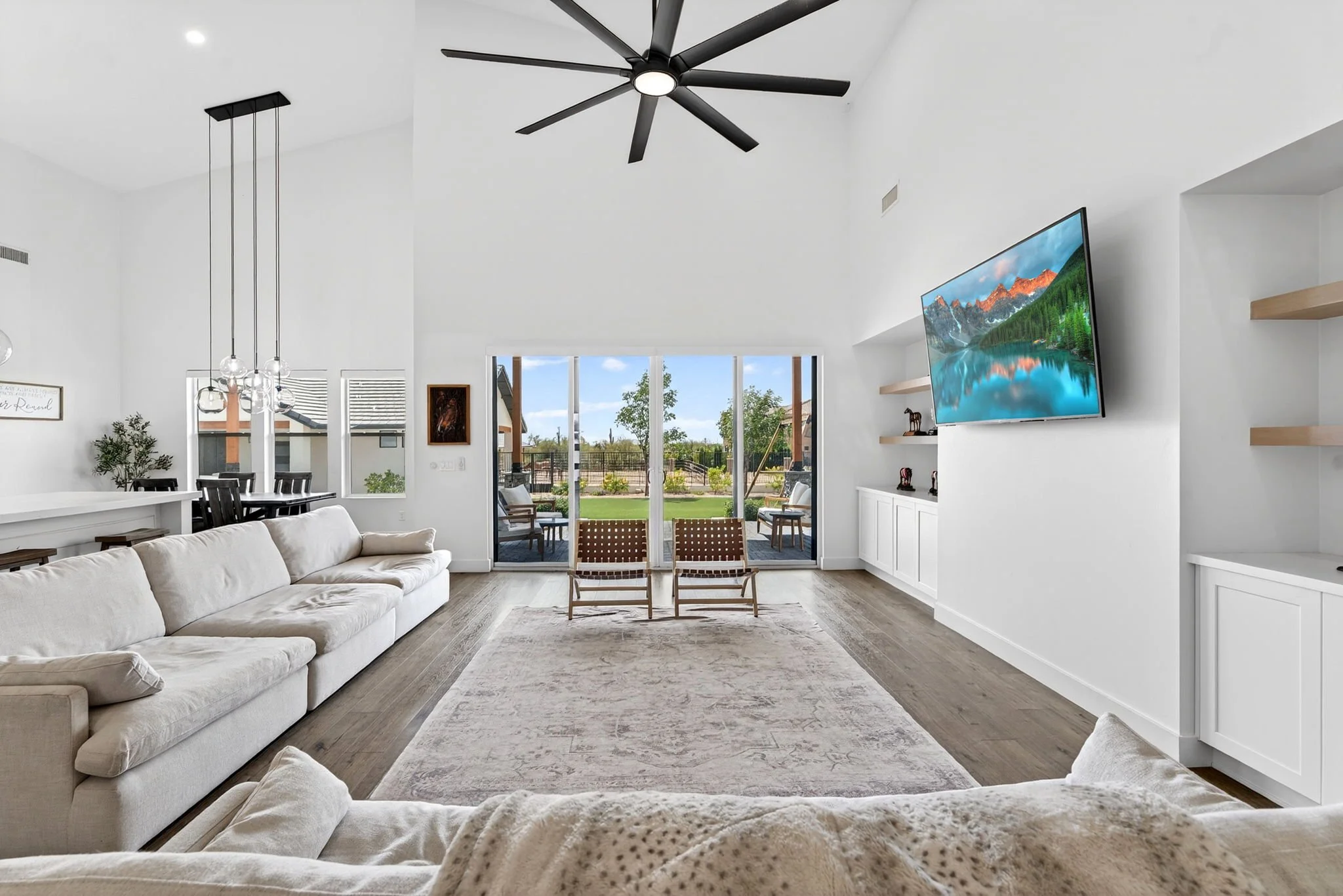 Modern living room with a white sectional sofa, a rug, a large window with a view of a backyard, a wall-mounted flat-screen TV, and floating shelves.