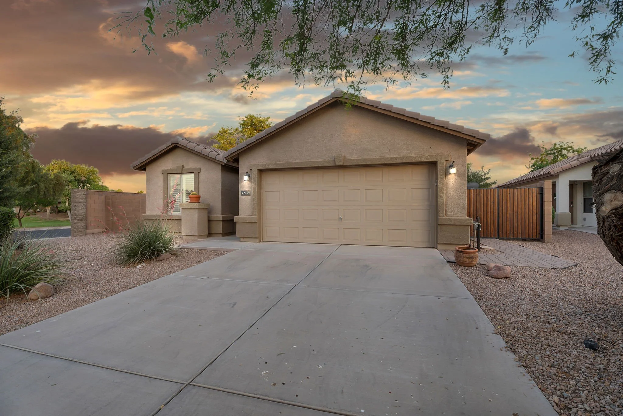 A residential house with a beige garage door, desert landscaping, and a concrete driveway at sunset. There are trees with green leaves and a cloudy sky in the background.