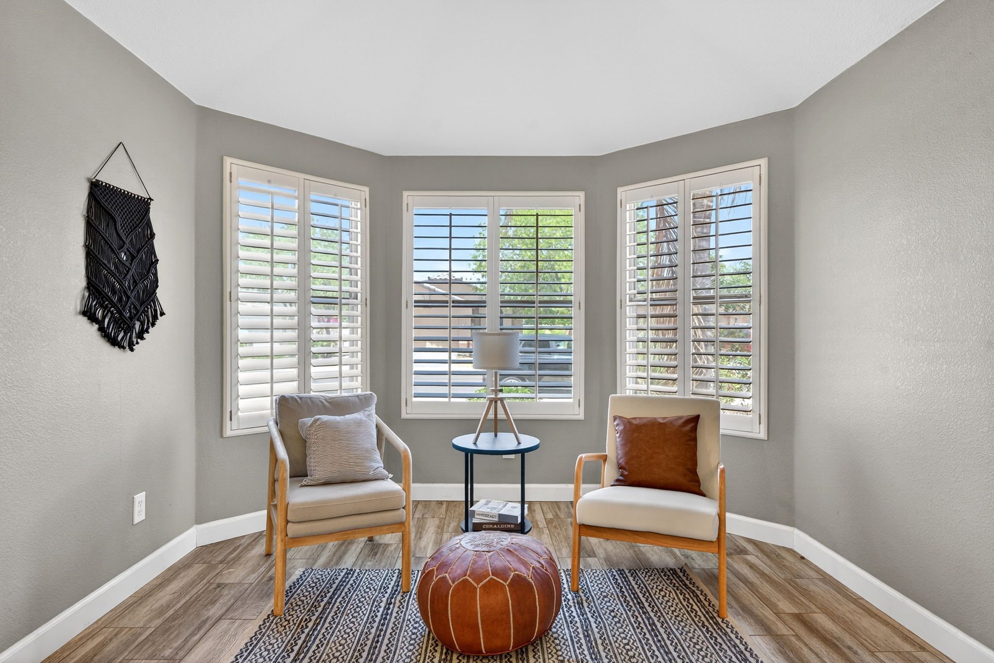 A cozy sitting room with two armchairs, a small side table with a lamp, and a leather pouf on a patterned rug, all lit by sunlight through large bay windows with white shutters.
