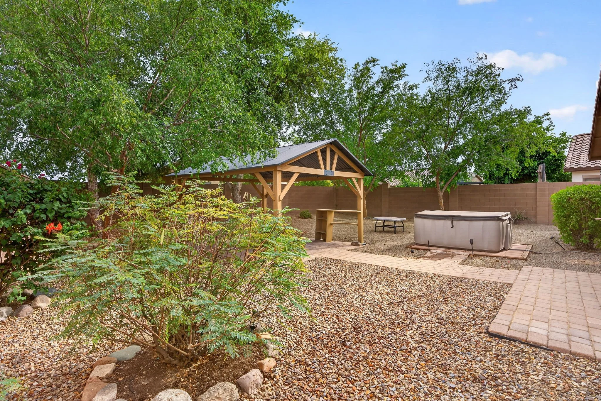 A backyard with large green trees, a wooden pergola with a roof, a small table underneath, a brick pathway, and a hot tub on a gravel surface, enclosed by a brown fence and neighboring house with a tile roof.
