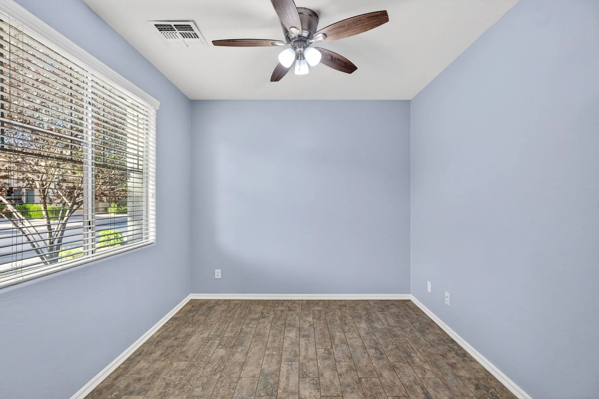 Empty room with light blue walls, a large window with blinds, a ceiling fan with lights, and wooden flooring.