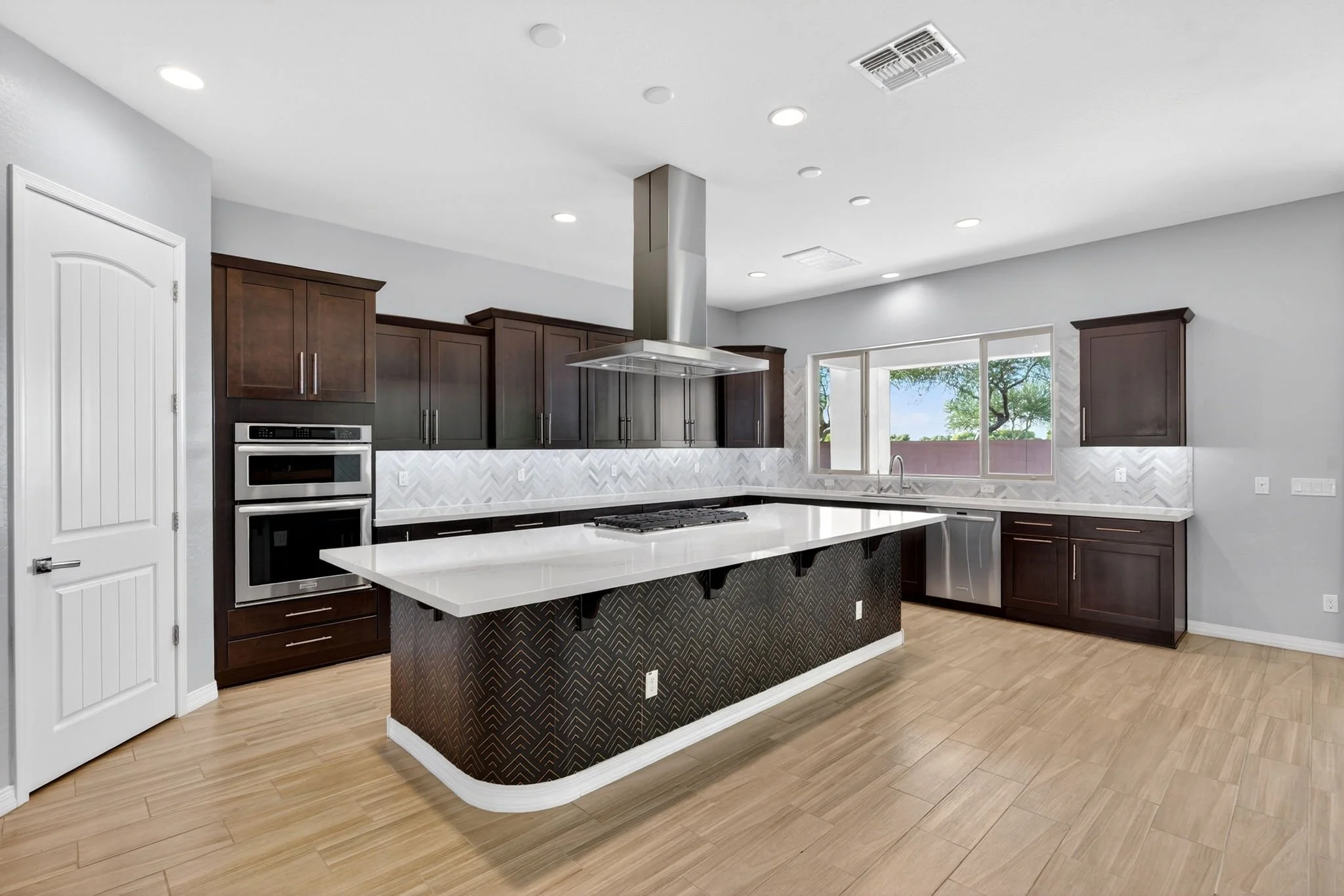 Modern kitchen with dark wood cabinets, white island with patterned black base, stainless steel stove and range hood, light wood flooring, white walls, large window with scenic view.