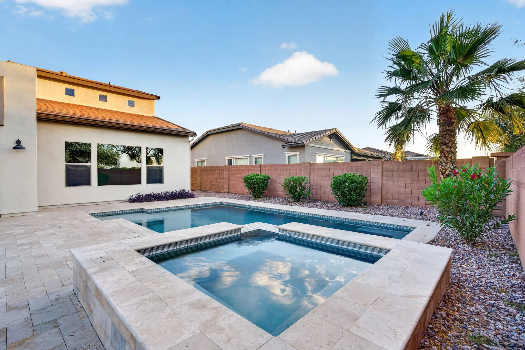 Backyard with a rectangular swimming pool, surrounded by light-colored stone paving, with bushes, a tall palm tree, a brick privacy wall, and houses beyond under a clear blue sky.