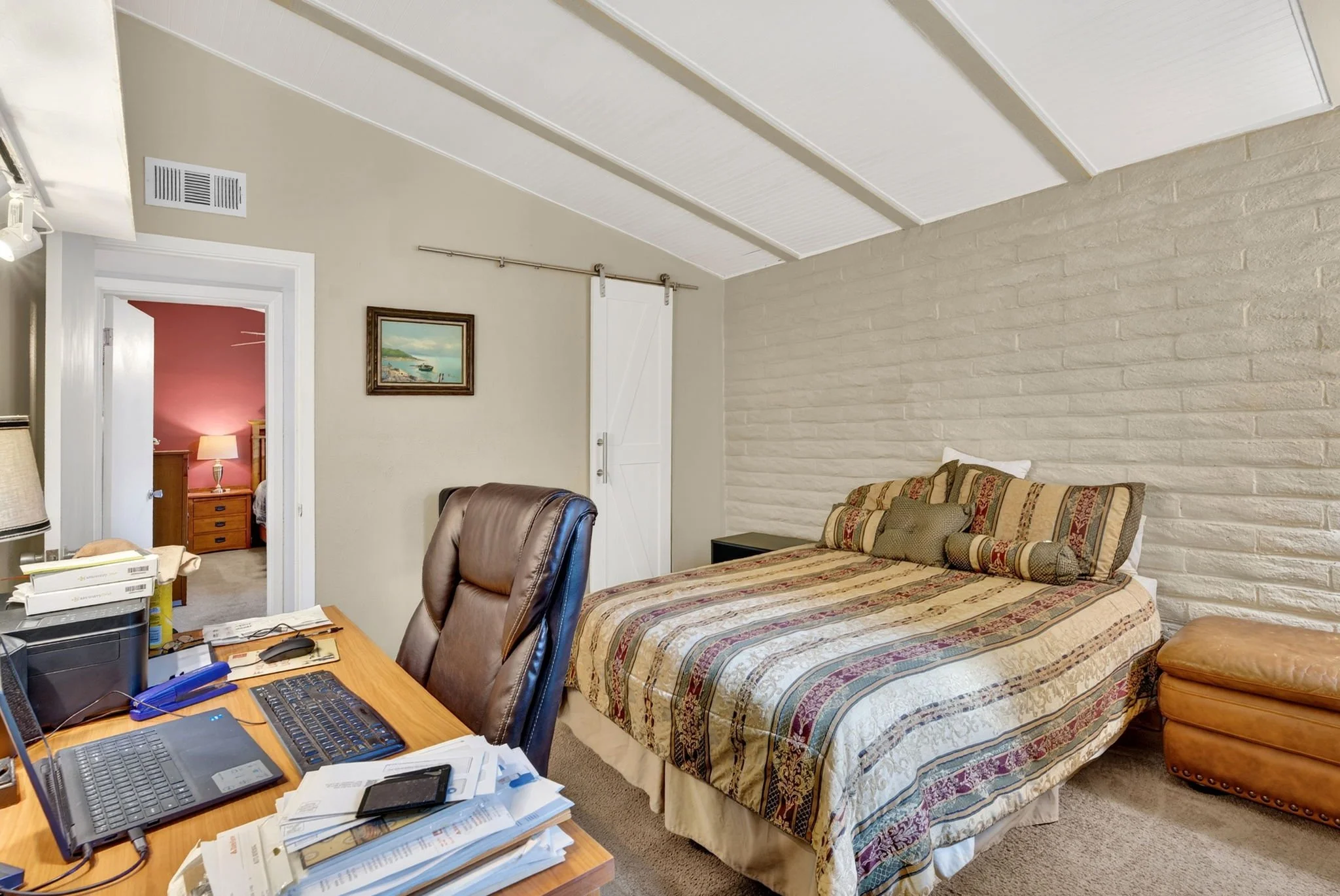 A cluttered bedroom with a bed and a workspace table. The bed has a patterned quilt and pillows. The room features a white brick accent wall, a framed painting, and a sliding barn door. The workspace includes a desk with papers, a computer, and offic