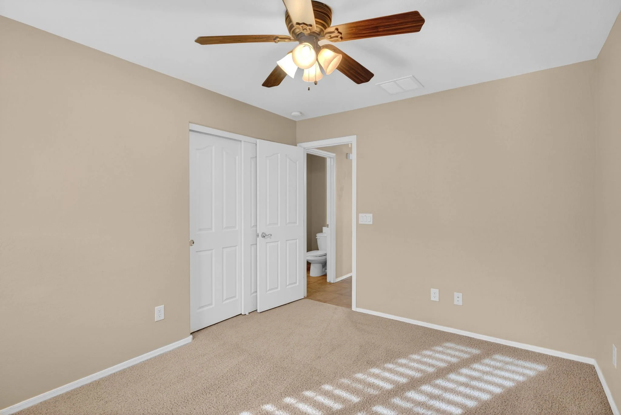 Empty room with beige walls, beige carpet, and a ceiling fan with lights. A door is open to a bathroom with a toilet visible.
