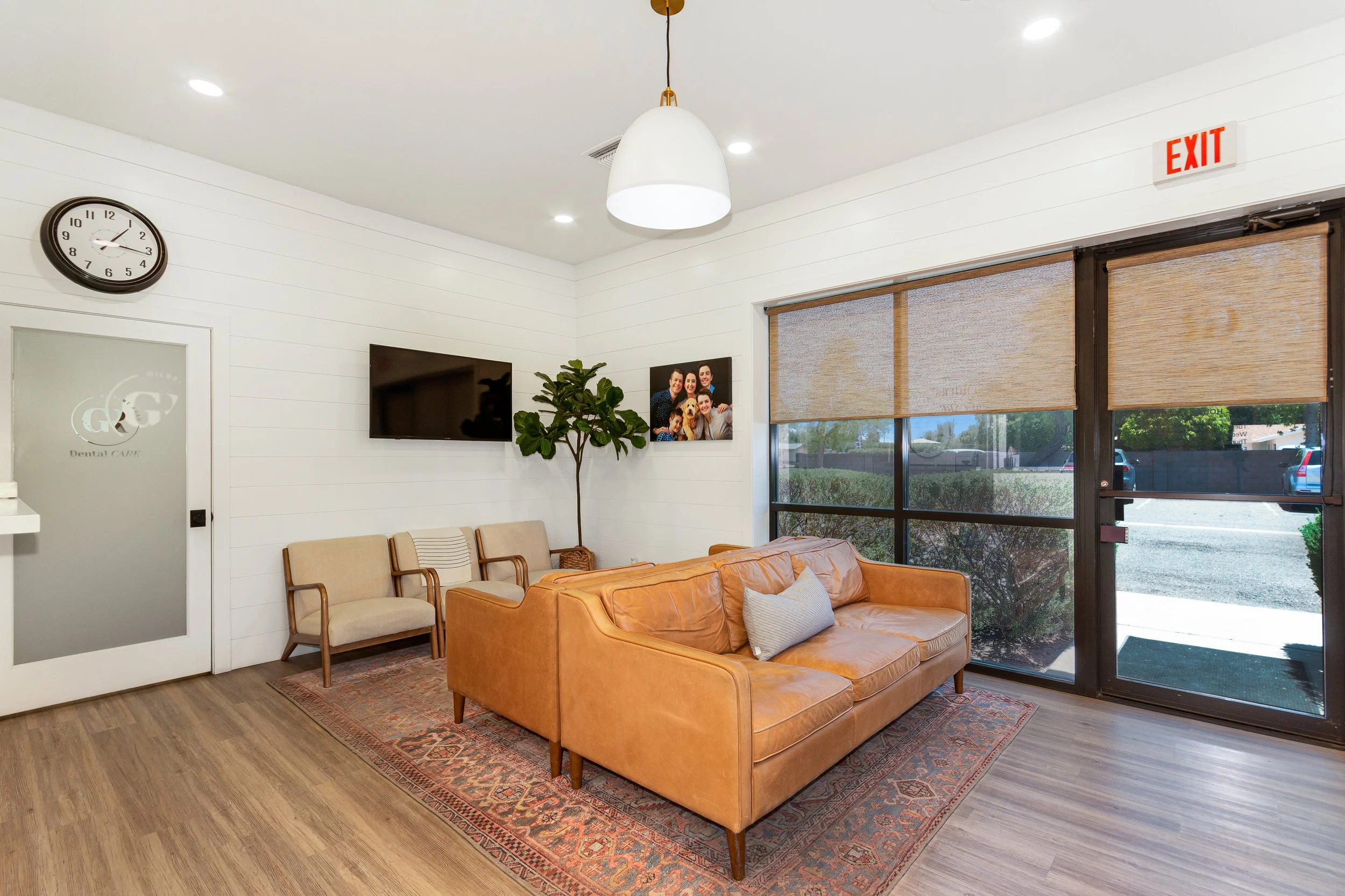 A waiting room with beige chairs, a leather sofa, a wall-mounted TV, a large window with bamboo blinds, a potted plant, and family photos on the wall.