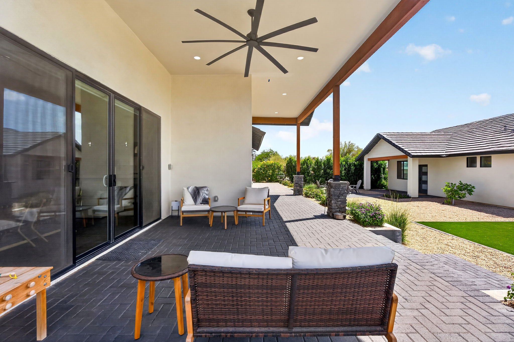 Covered outdoor patio with black pavers, white ceiling with ceiling fan, glass sliding door, outdoor furniture including a wicker chair with cushions, small round table, and another white cushioned chair, overlooking a landscaped yard with a small bu