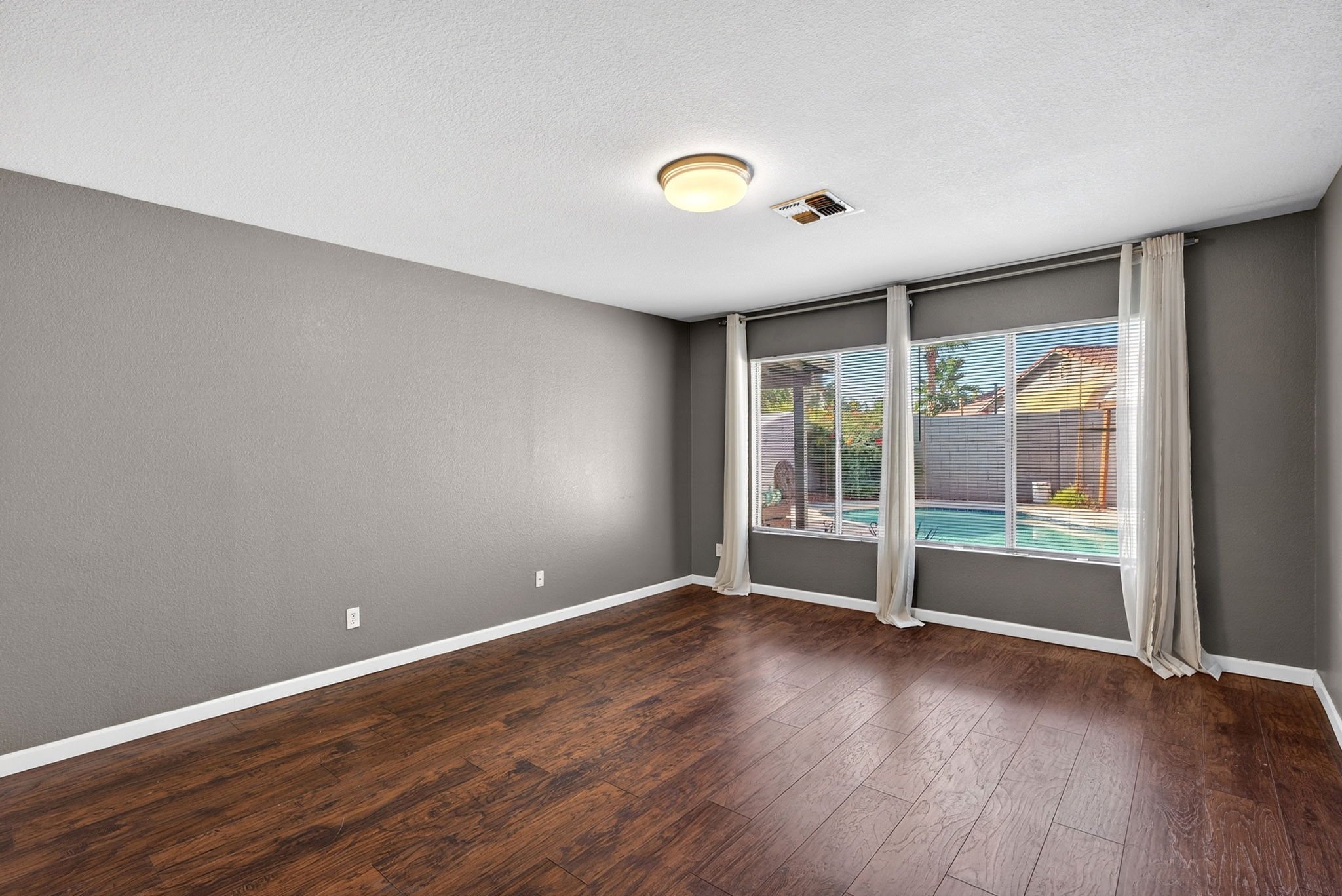 Empty living room with gray walls, wooden flooring, large window with white curtains, and ceiling light fixture.