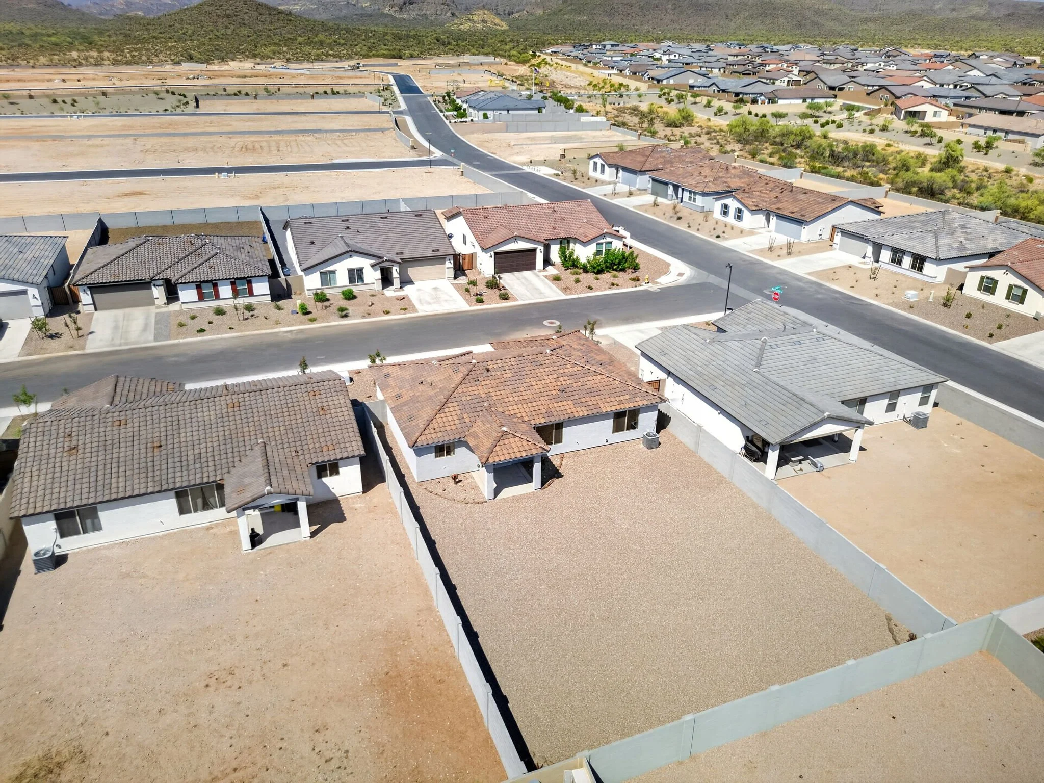 Aerial view of a suburban neighborhood with houses featuring tiled and shingle roofs, some with yards, and a large empty lot under development. In the background, there are hills or mountains.