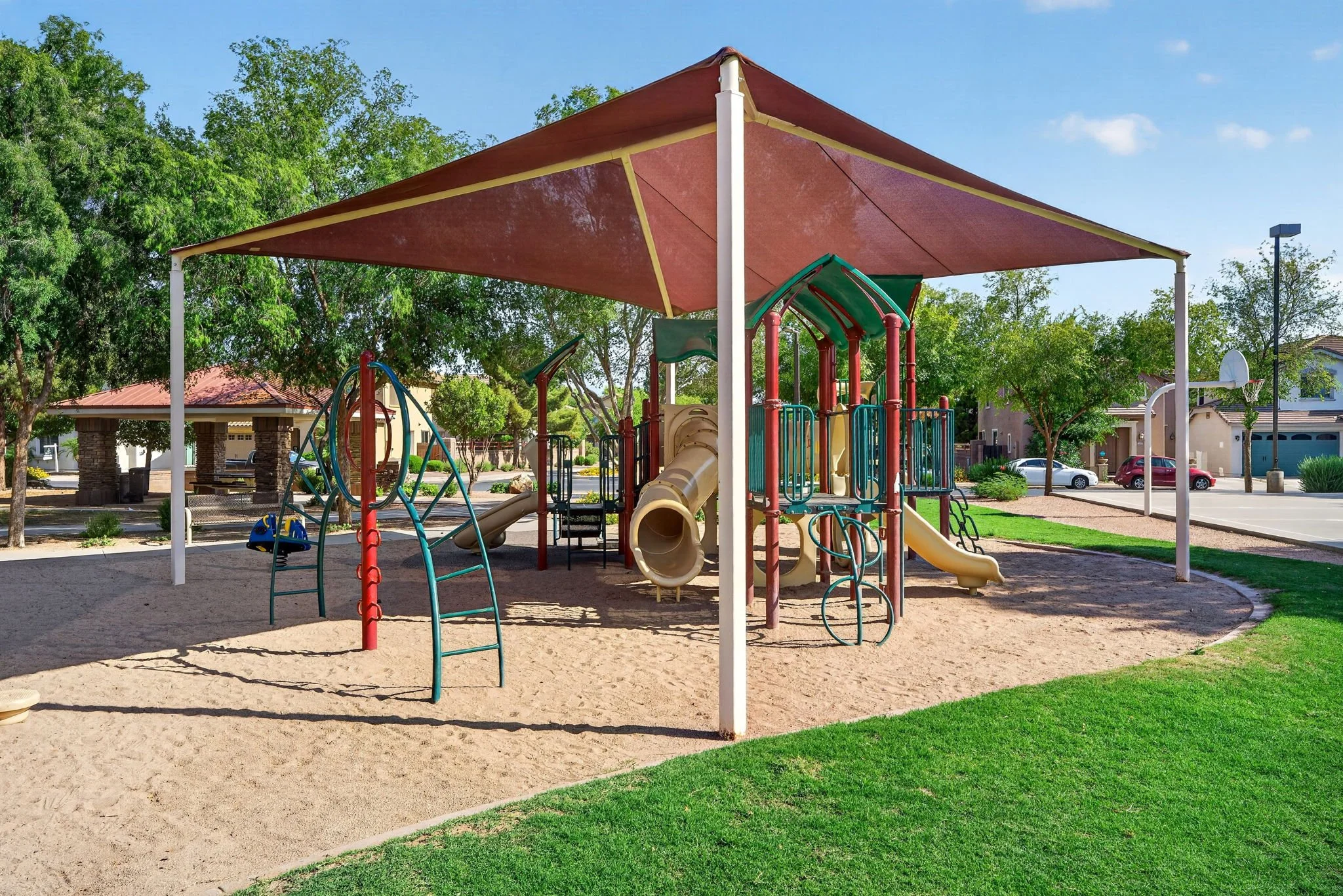 Colorful playground equipment under a shade canopy in a park with trees, grass, and a parking lot in the background.