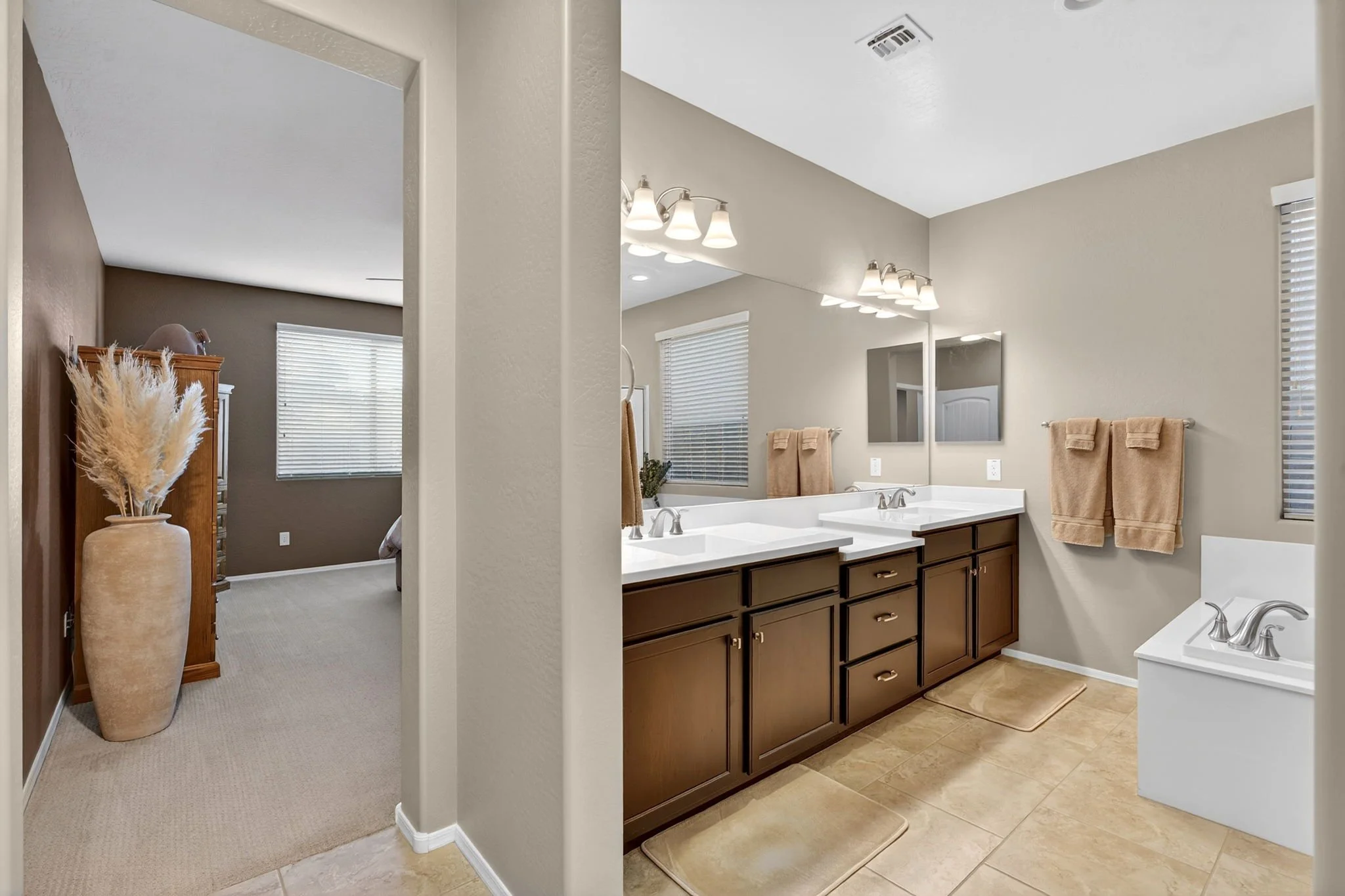 Bathroom with dual sinks, brown cabinets, and beige towels on wall. Adjacent bedroom with window, carpet, and tall vase with pampas grass.