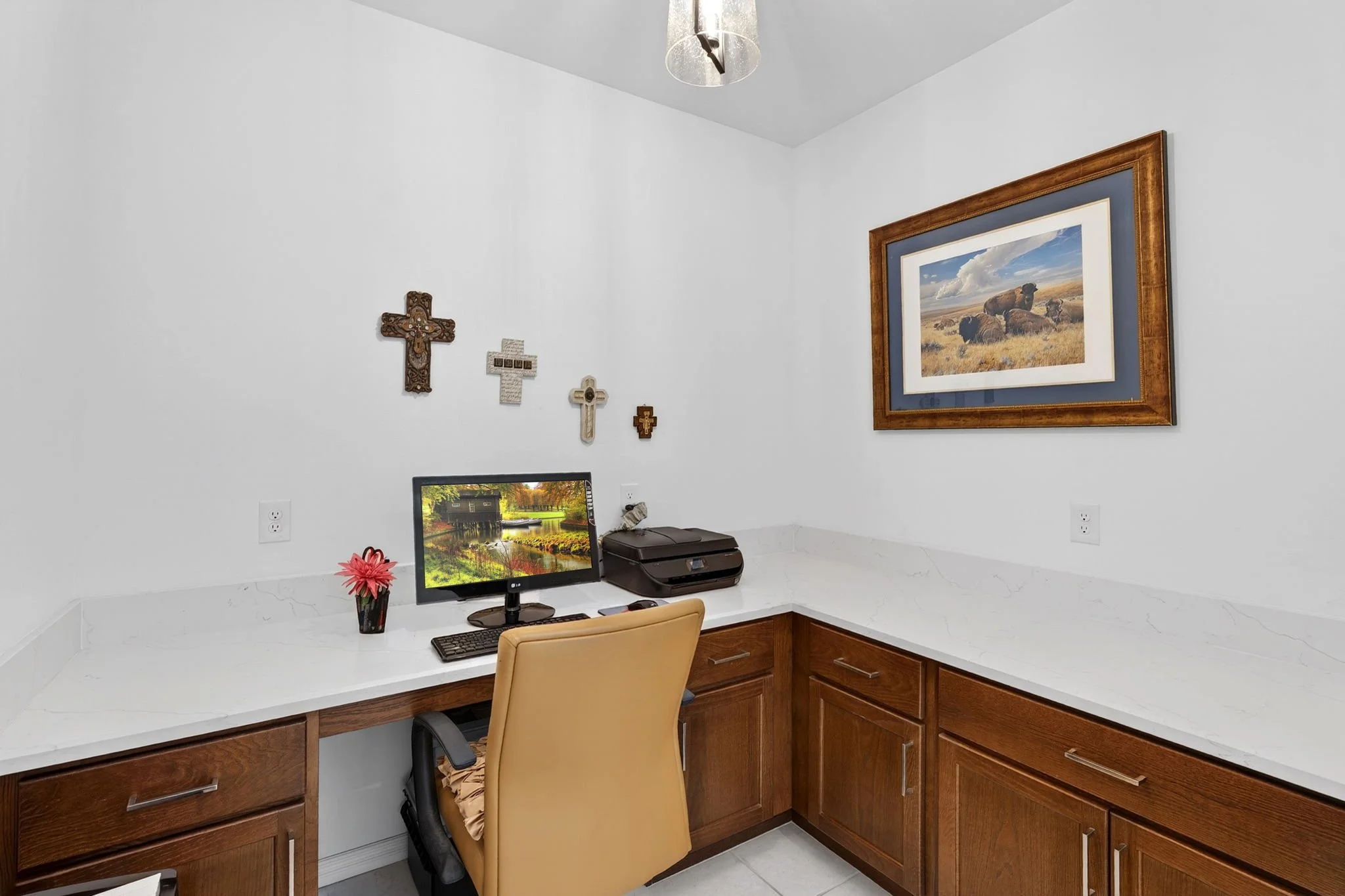 A corner of a home office with a white L-shaped desk, a computer monitor, a beige office chair, and wall decorations including framed pictures and wooden crosses.