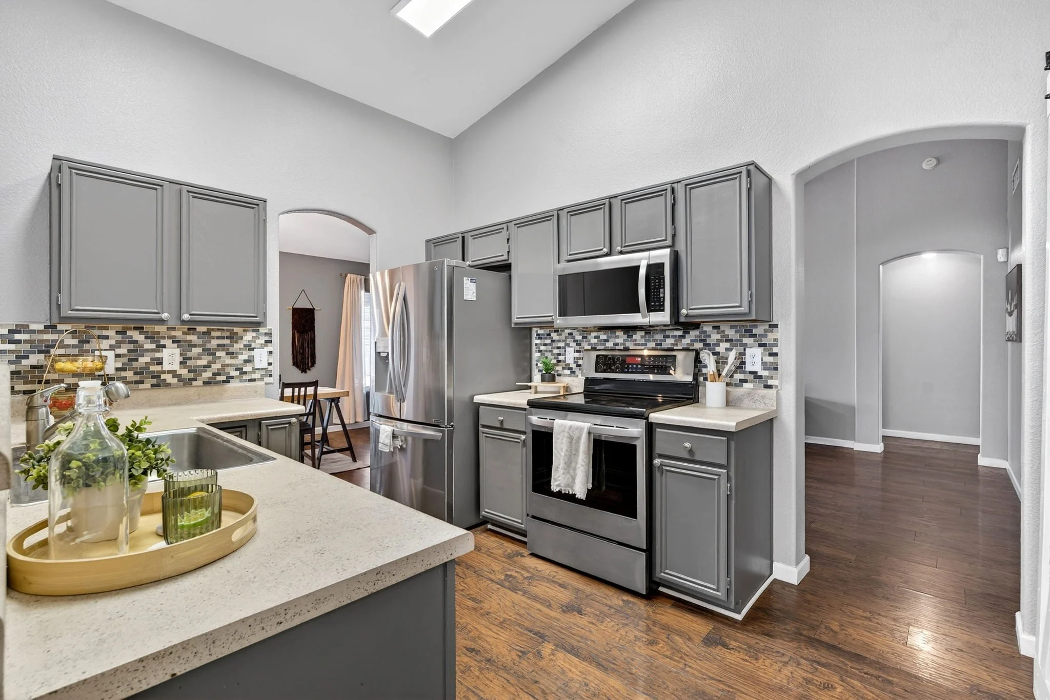 A modern kitchen with gray cabinets, stainless steel appliances, a mosaic tile backsplash, wooden floors, and a ceiling skylight.