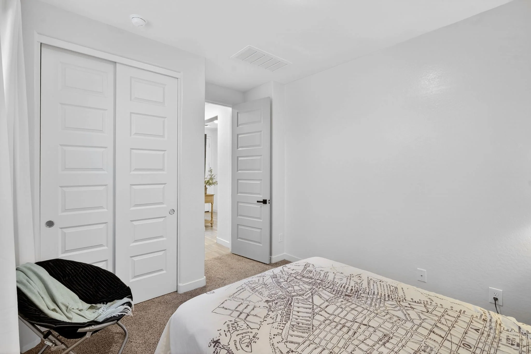 Bedroom with white walls, bed with city map design bedspread, hikc chair, partially open closet doors, ceiling vent, and hallway leading to another room with a table and plant.