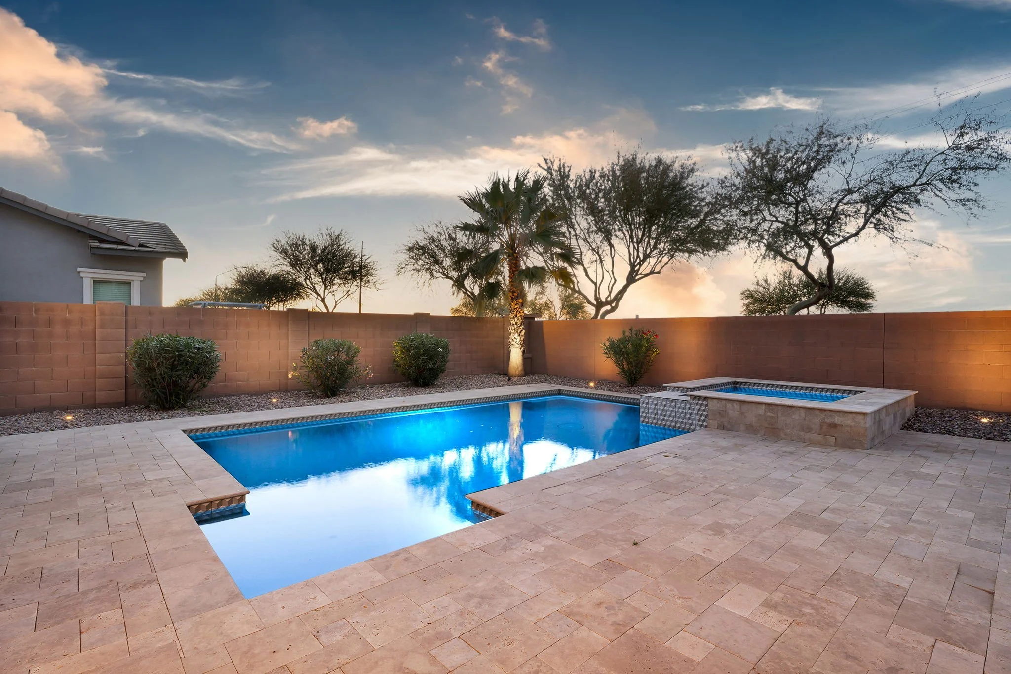 Backyard with swimming pool, palm trees, and other trees, enclosed by a brick wall, during sunset or dusk.