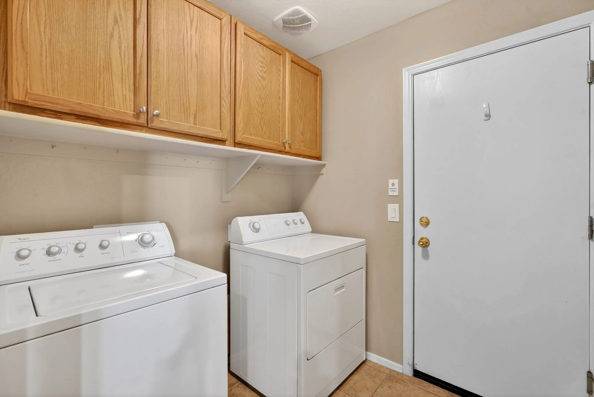 Laundry room with white washer and dryer, wooden cabinets above, beige walls, and a white door with a gold doorknob.