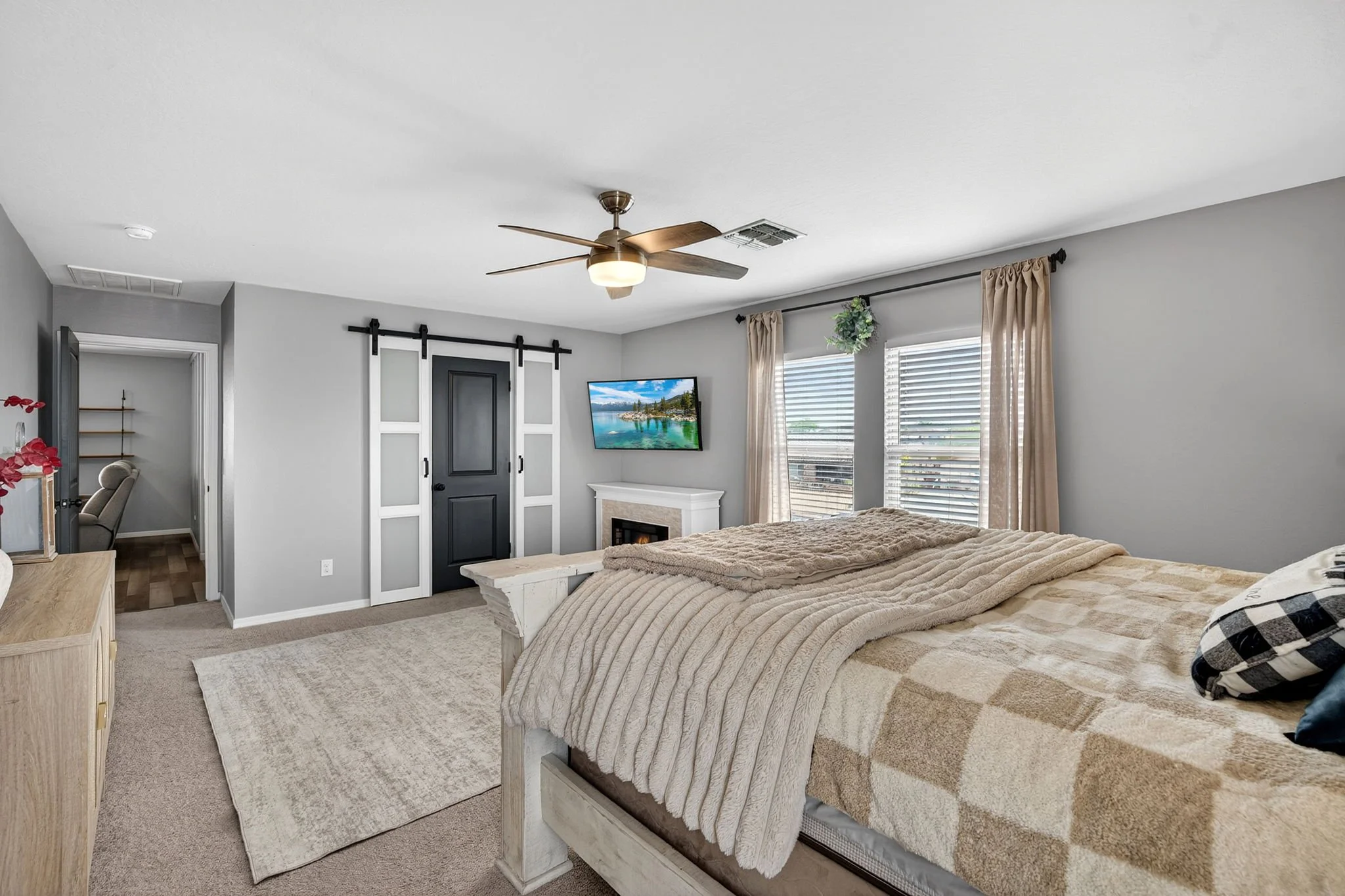 A bedroom with a white ceiling and walls, a ceiling fan, a window with beige curtains, a bed with a beige blanket and a checkered pillow, a wall-mounted TV, and sliding closet doors.