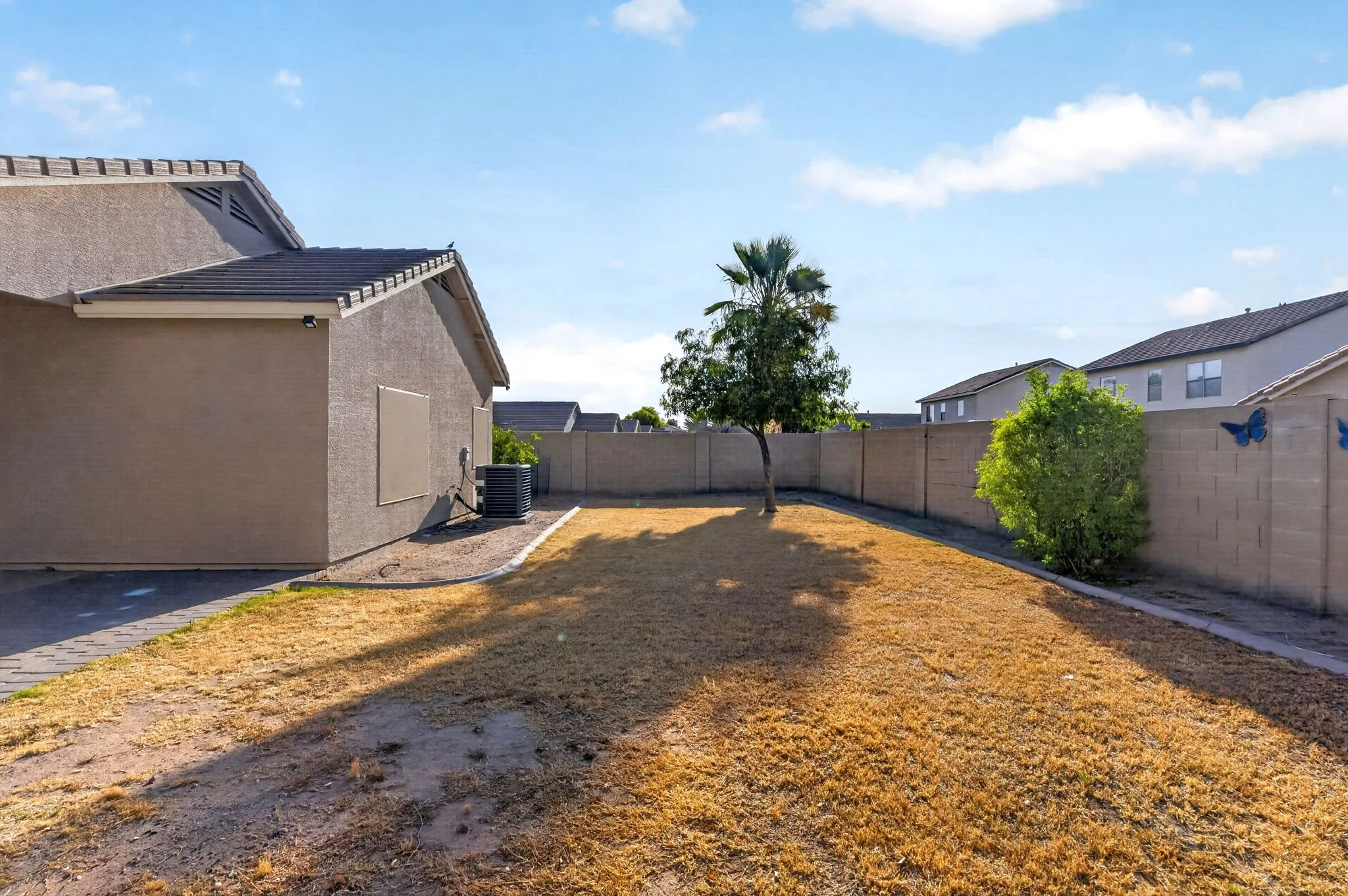 A backyard with a house, a tree, a tall beige fence, and a butterfly decoration on the fence, under a blue sky with some clouds.
