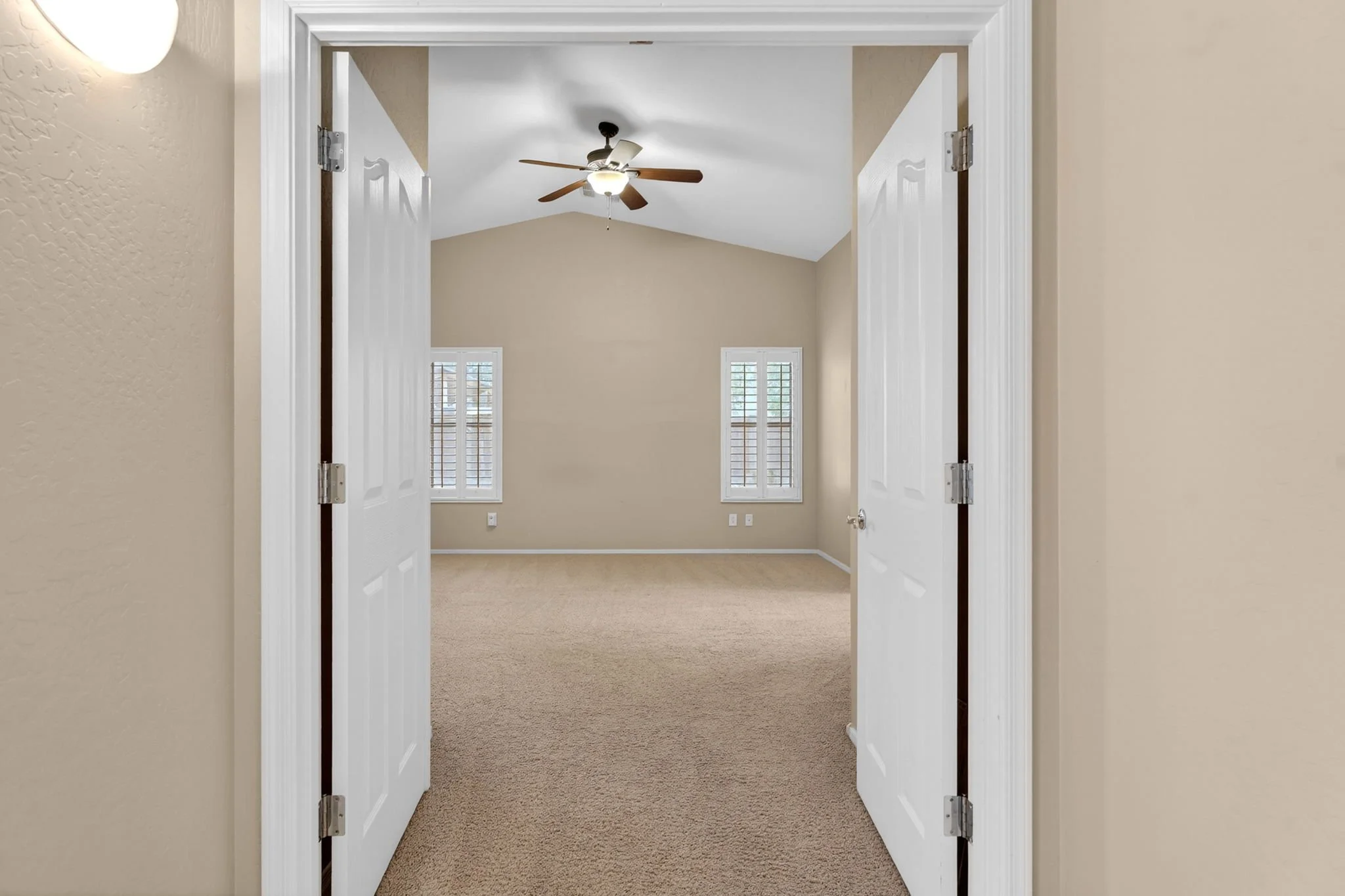 View through double white doors into an empty room with beige walls, two windows with shutters, and a ceiling fan with light in the center of the ceiling.