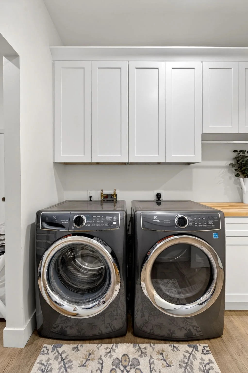 A laundry room with gray front-loading washing machine and dryer, white cabinets above, wooden countertop on the right, and a potted plant to the side.