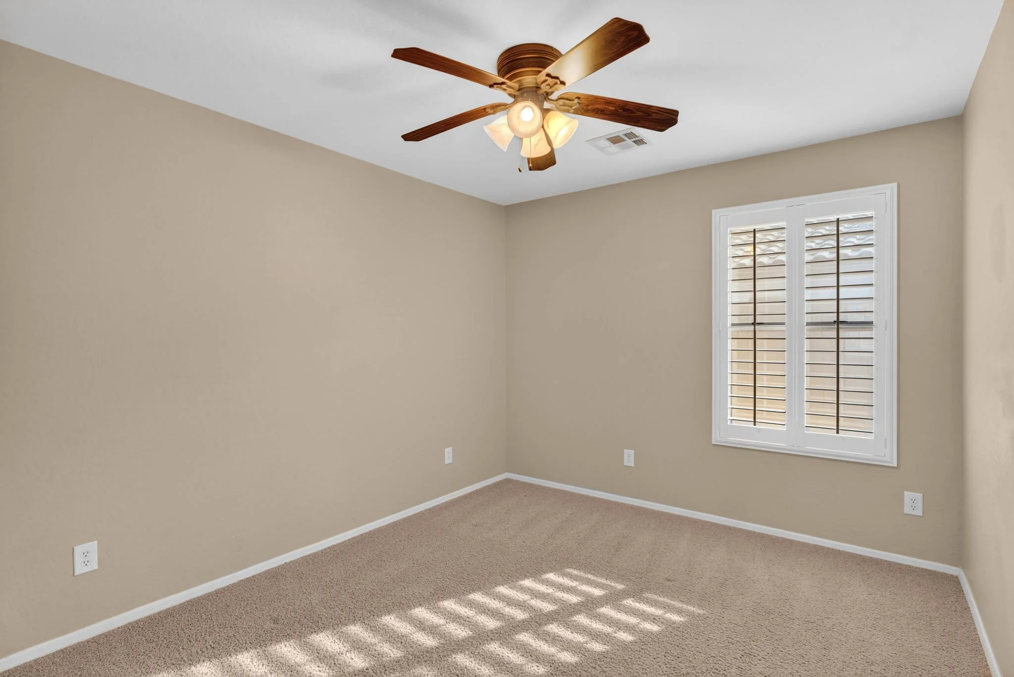 An empty beige room with carpeted floor, a window with shutters, a ceiling fan with lights, and electrical outlets.