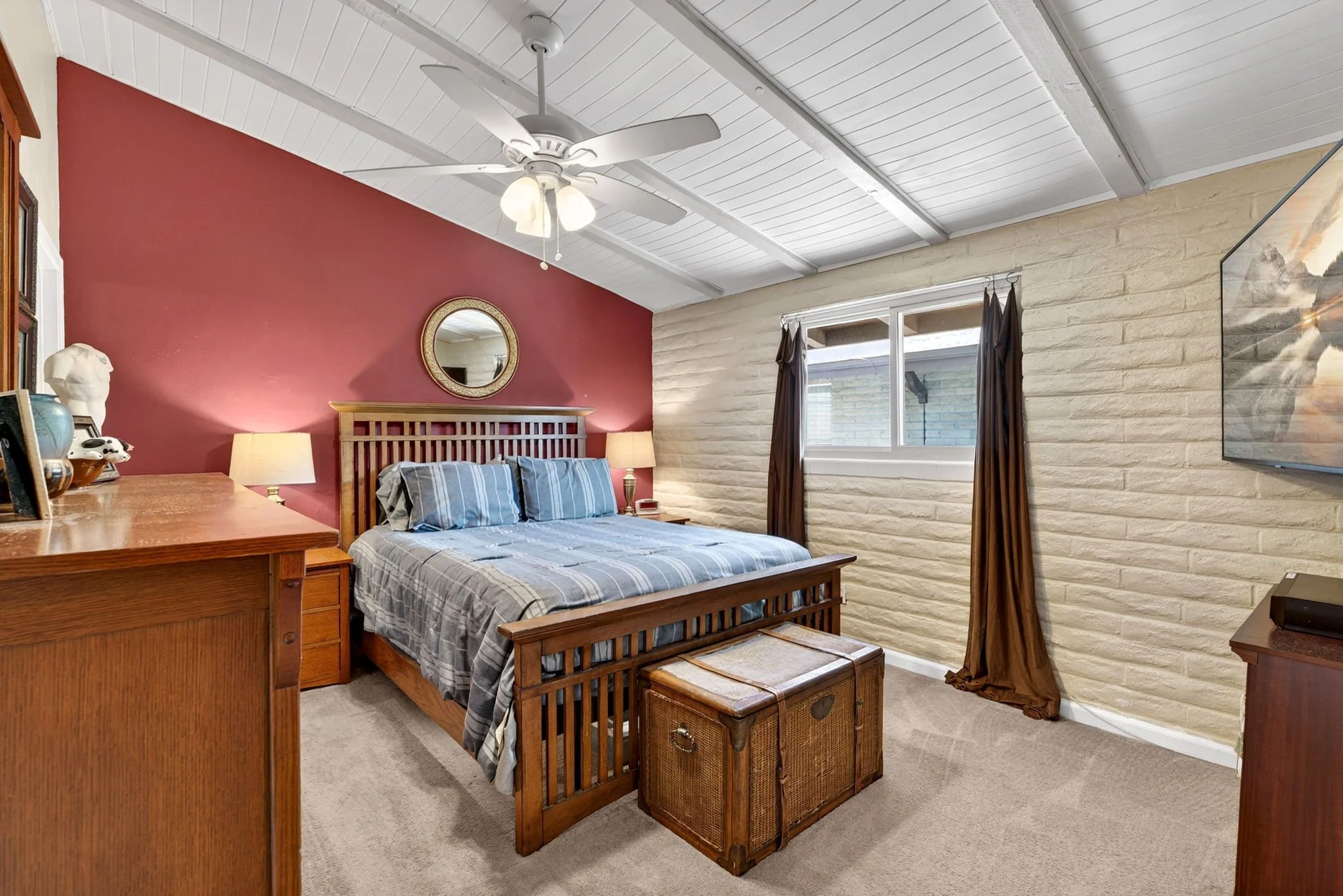 A bedroom with a wooden bed frame and a striped bedspread, two nightstands with lamps, a window with dark curtains, a ceiling fan with lights, a red accent wall, and a white brick wall.