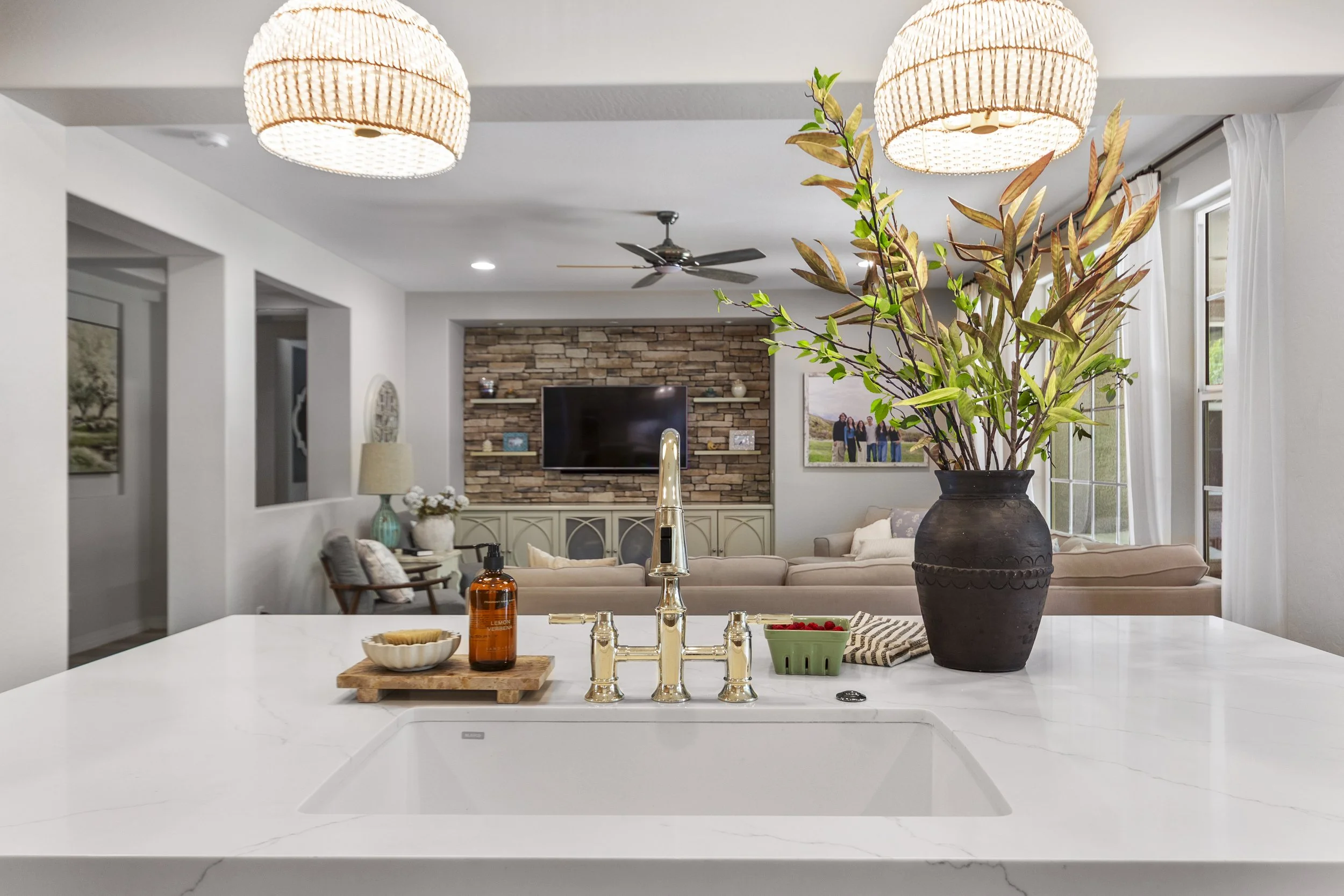 A modern kitchen with a white island countertop, a large black vase with green and brown foliage, a soap dispenser, a small bowl, and a green container. The background shows a living room with a stone accent wall, a flat-screen TV, picture frames, a 