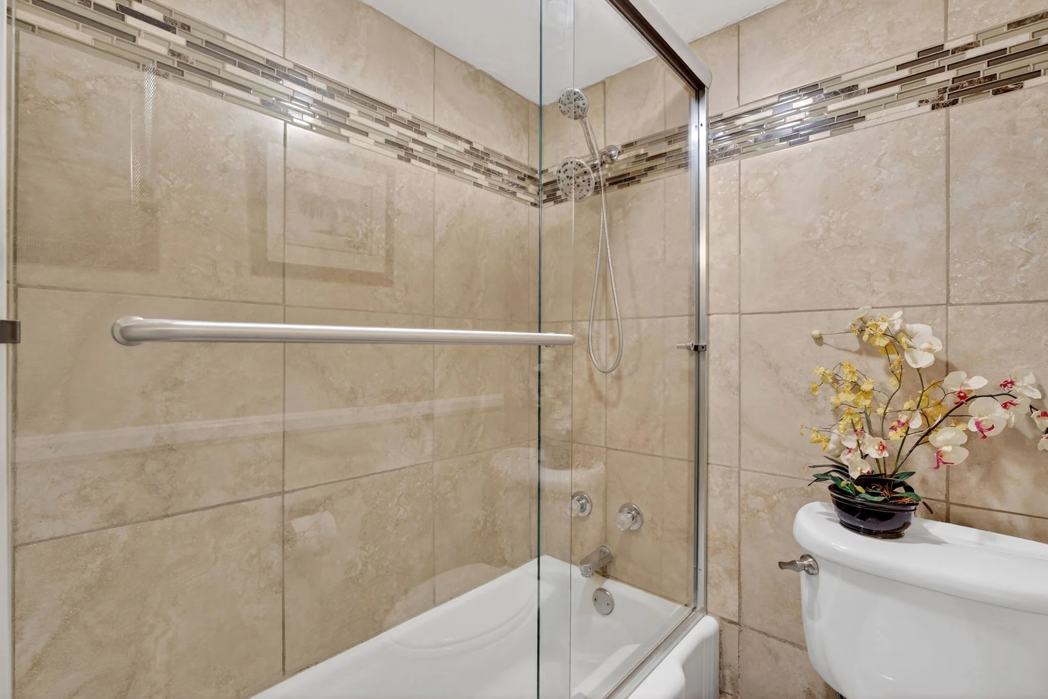 A clean bathroom with beige tiled walls and a shower area enclosed by glass doors. There is a decorative horizontal border of small glass tiles in brown and gray hues. Inside the shower, there is a handheld showerhead and control knobs. A white toile