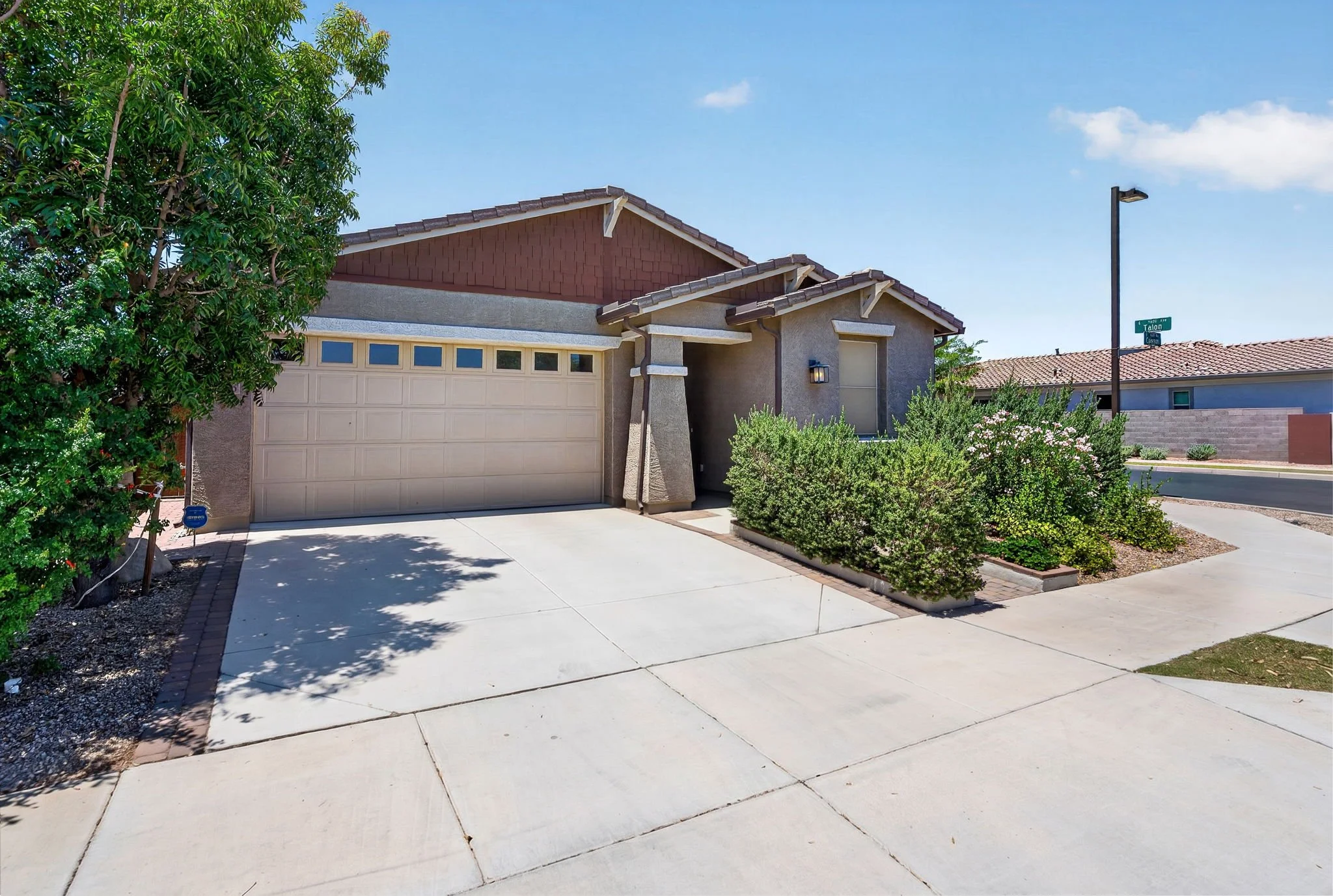 A house with a beige garage door, gray exterior walls, and a red-brown tiled roof. There are shrubs and a large tree in the front yard, and a street sign indicating the corner of Talon and Cason. The sky is clear with a few small clouds.