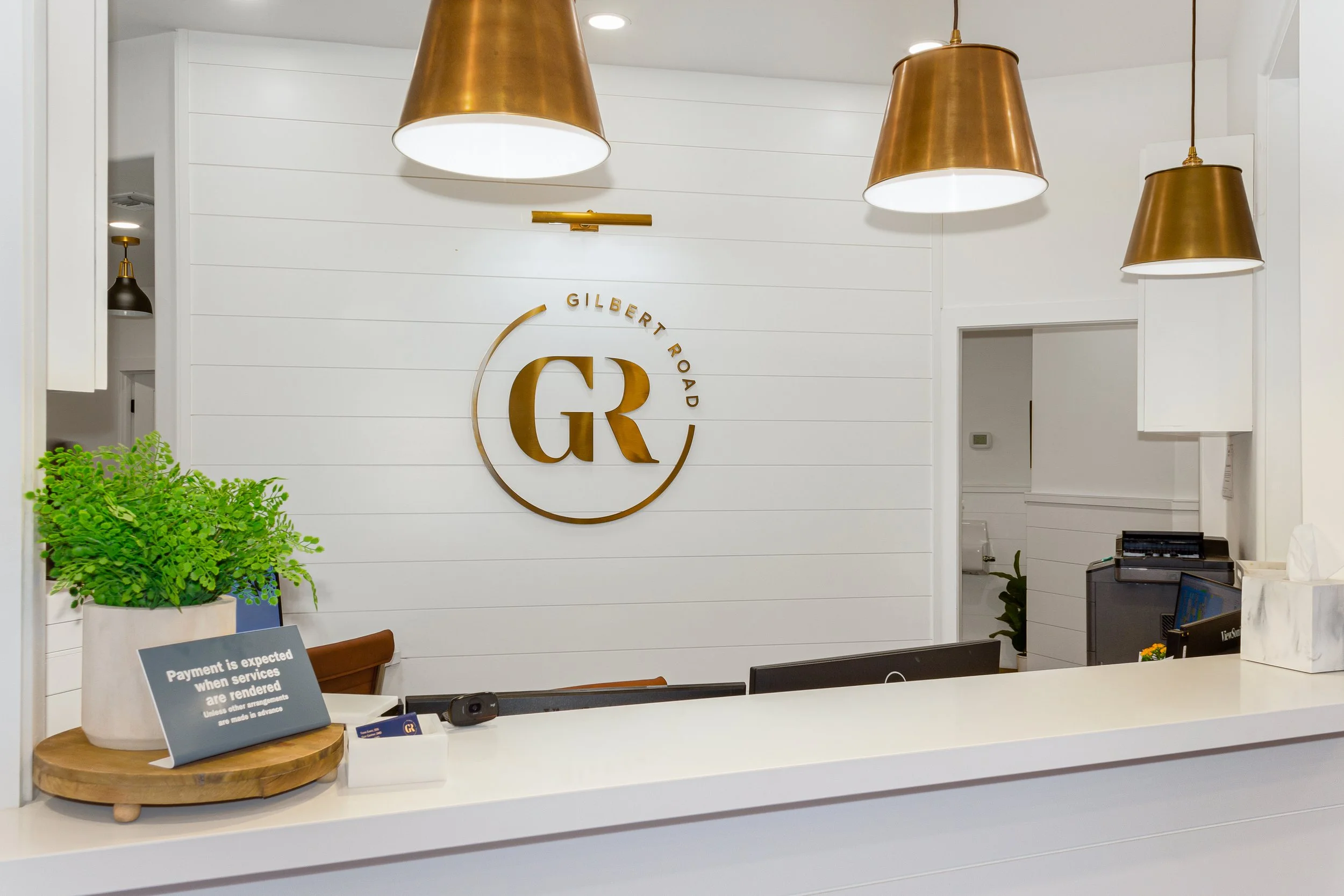 Interior of a Gilbert Road office or reception area with modern decor, including white walls, hanging pendant lights, a green plant on the counter, and a sign about payment expectations.