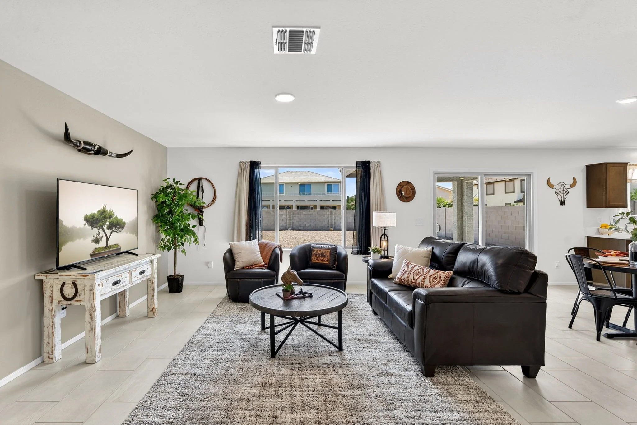 Open-concept living room with beige walls, a black leather sofa, two black armchairs, a white distressed TV stand with a TV, a round coffee table, a large window with blue and cream curtains, a potted plant, and various wall decor including a bull sk
