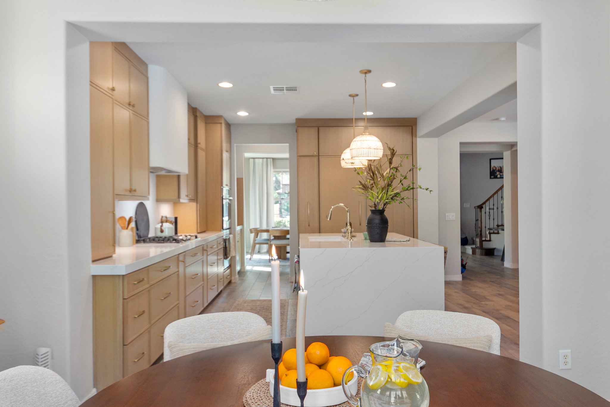 Interior view of a modern kitchen with wooden cabinets, white countertops, a kitchen island, hanging pendant lights, a vase with branches on the island, and a dining table with oranges, lemons, and candles.