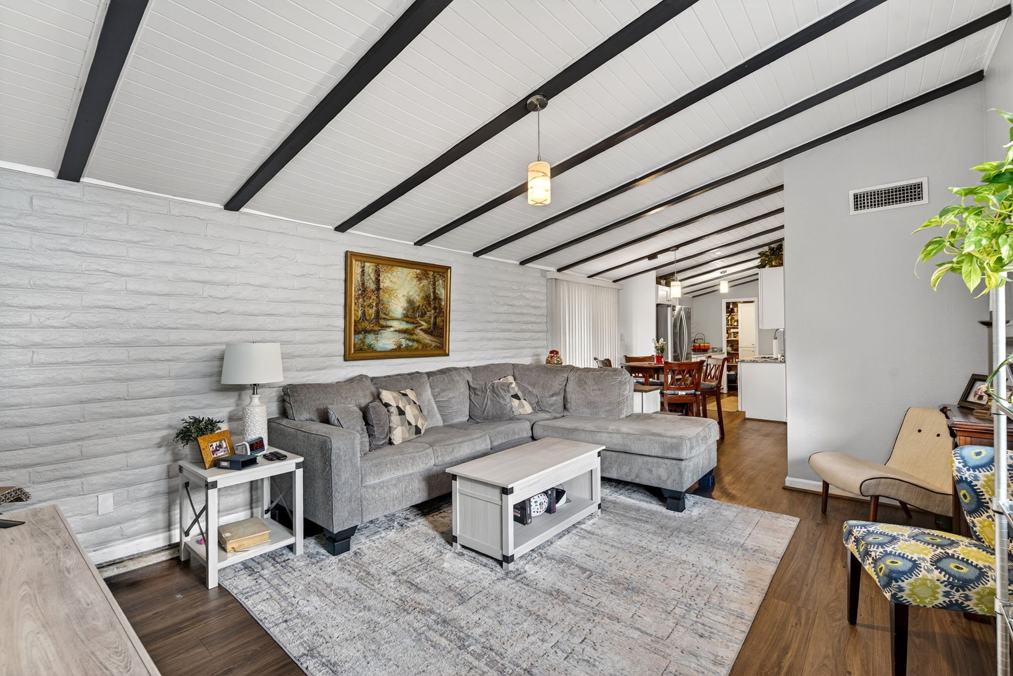 Living room with white brick wall, gray sectional sofa, wooden floor, and ceiling with black beams and hanging lights. There is a framed landscape painting on the wall, a white side table with picture frames, and a patterned armchair. Part of the kit