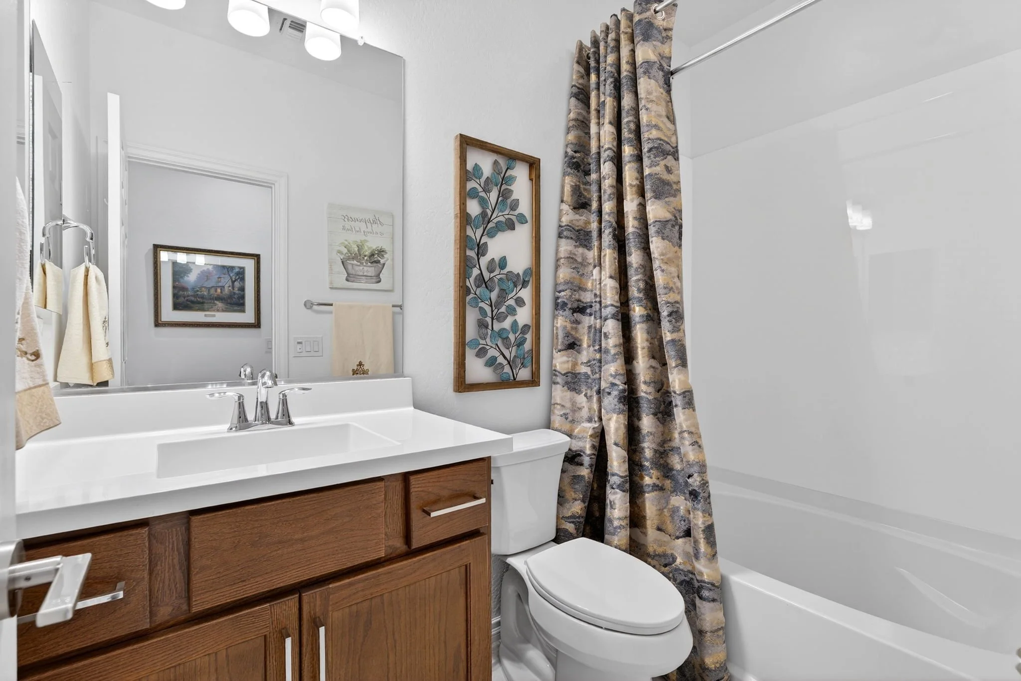 Bathroom with a white sink vanity, a mirror, framed artwork, a mirror above the sink, beige towels, a shower with patterned curtains, and a toilet.