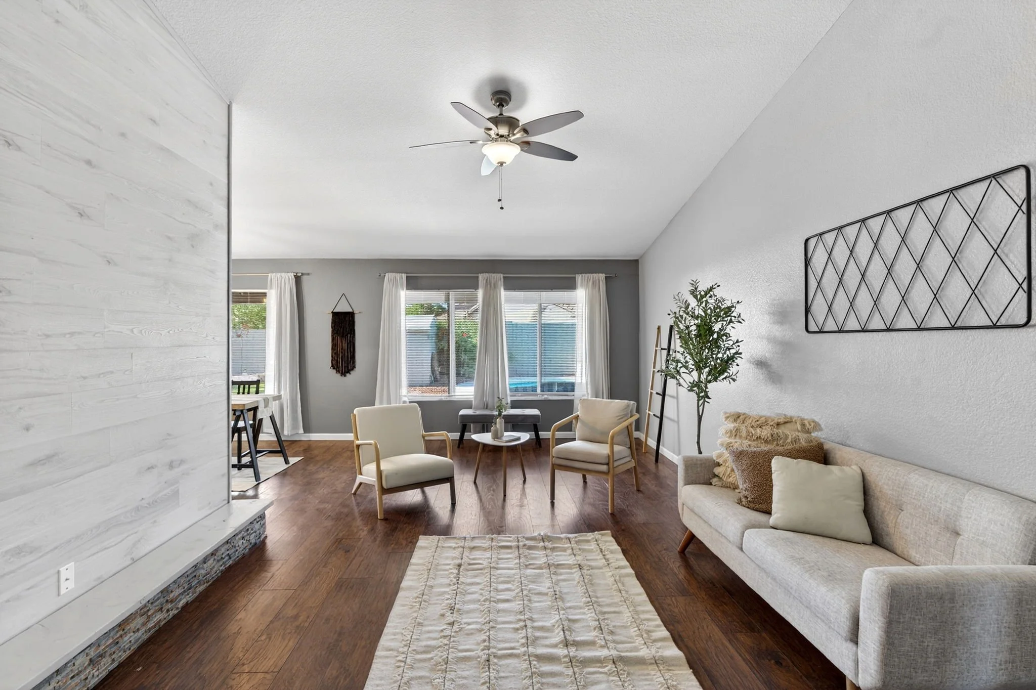 Interior of a living room with white walls, large windows with white curtains, a ceiling fan, and modern furniture including a beige sofa, chairs, and a small coffee table. Decorative items and a lush green plant are also visible.