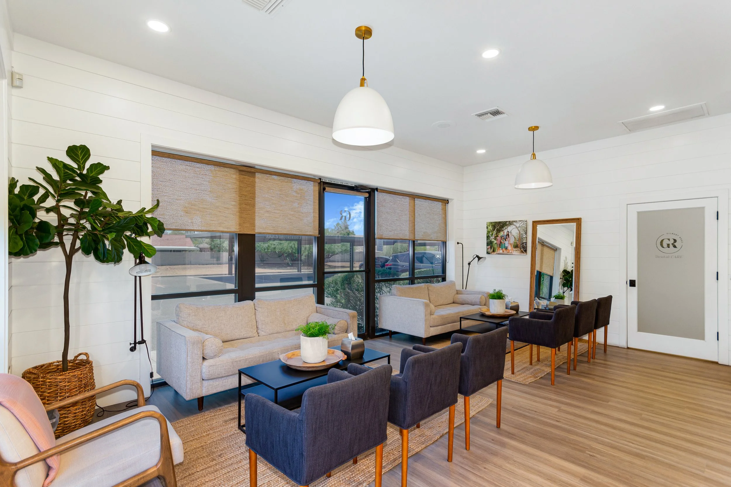 A modern waiting room with beige and dark gray armchairs arranged around black coffee tables, large windows with beige shades, white paneled walls, ceiling lights, green plants, and artwork.