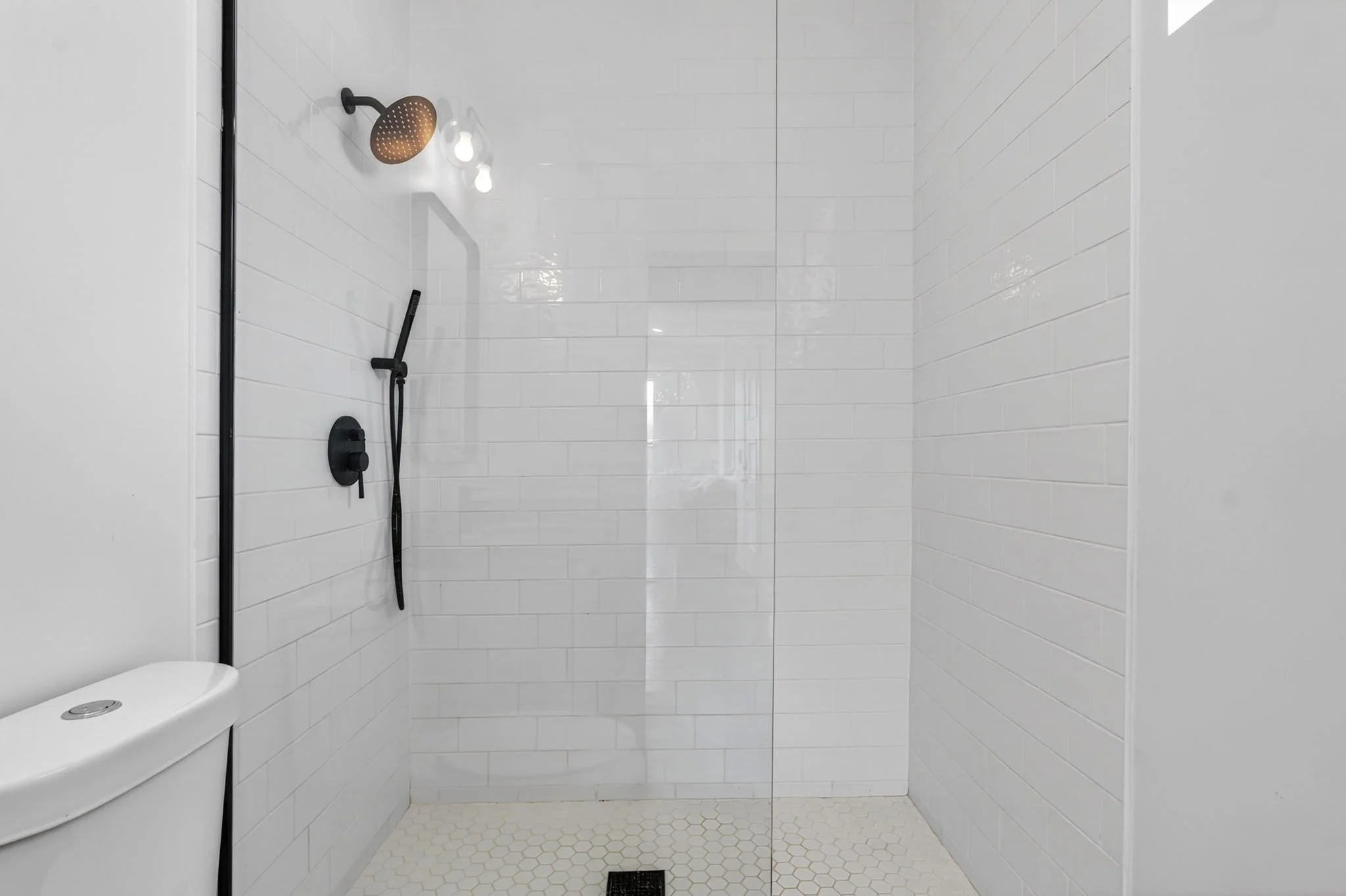 Shower stall with white tile walls, a brown showerhead, a handheld shower wand, a small white corner ledge, and a drain on the cream hexagon-tile floor.