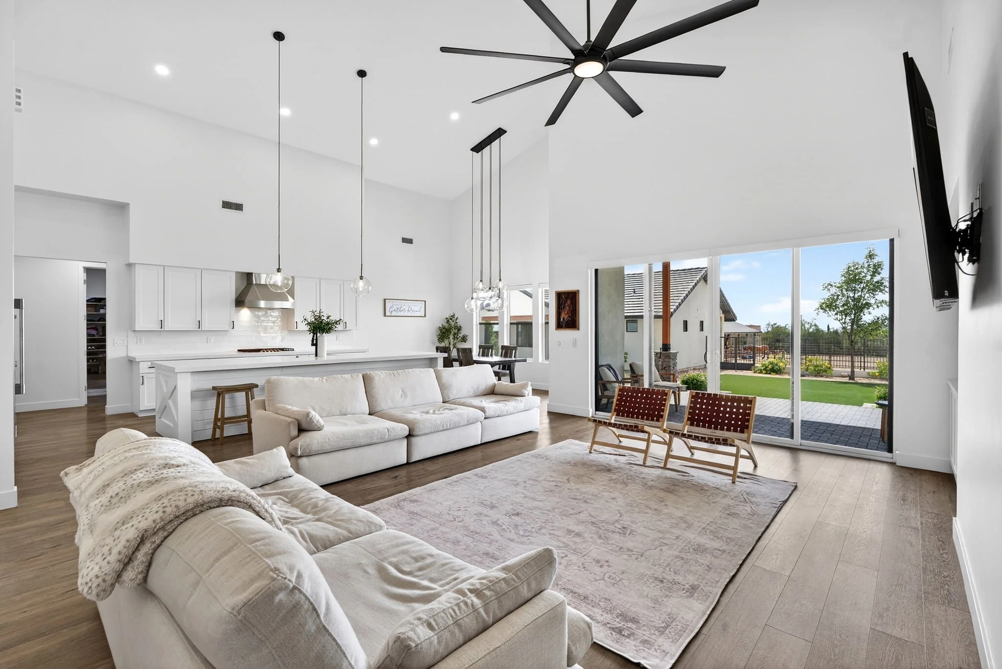 Living room with white couches, cream throw blanket, area rug, natural wood flooring, large sliding glass doors leading to backyard, ceiling fan, pendant lighting, kitchen in background with white cabinets, stainless steel range hood, and dining area