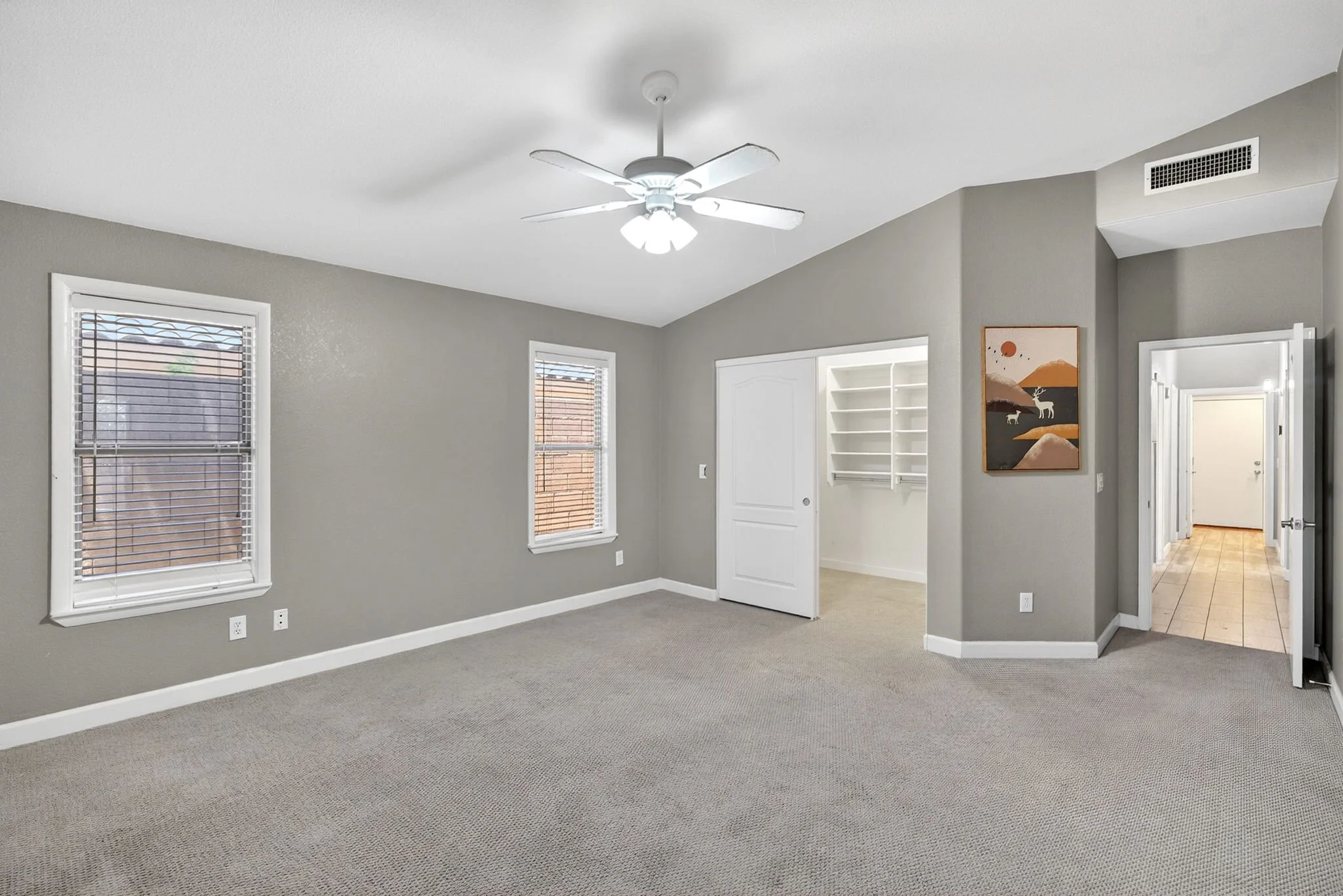 Empty room with two windows, gray walls, carpeted floor, ceiling fan, and an artwork featuring a mountain scene with deer.
