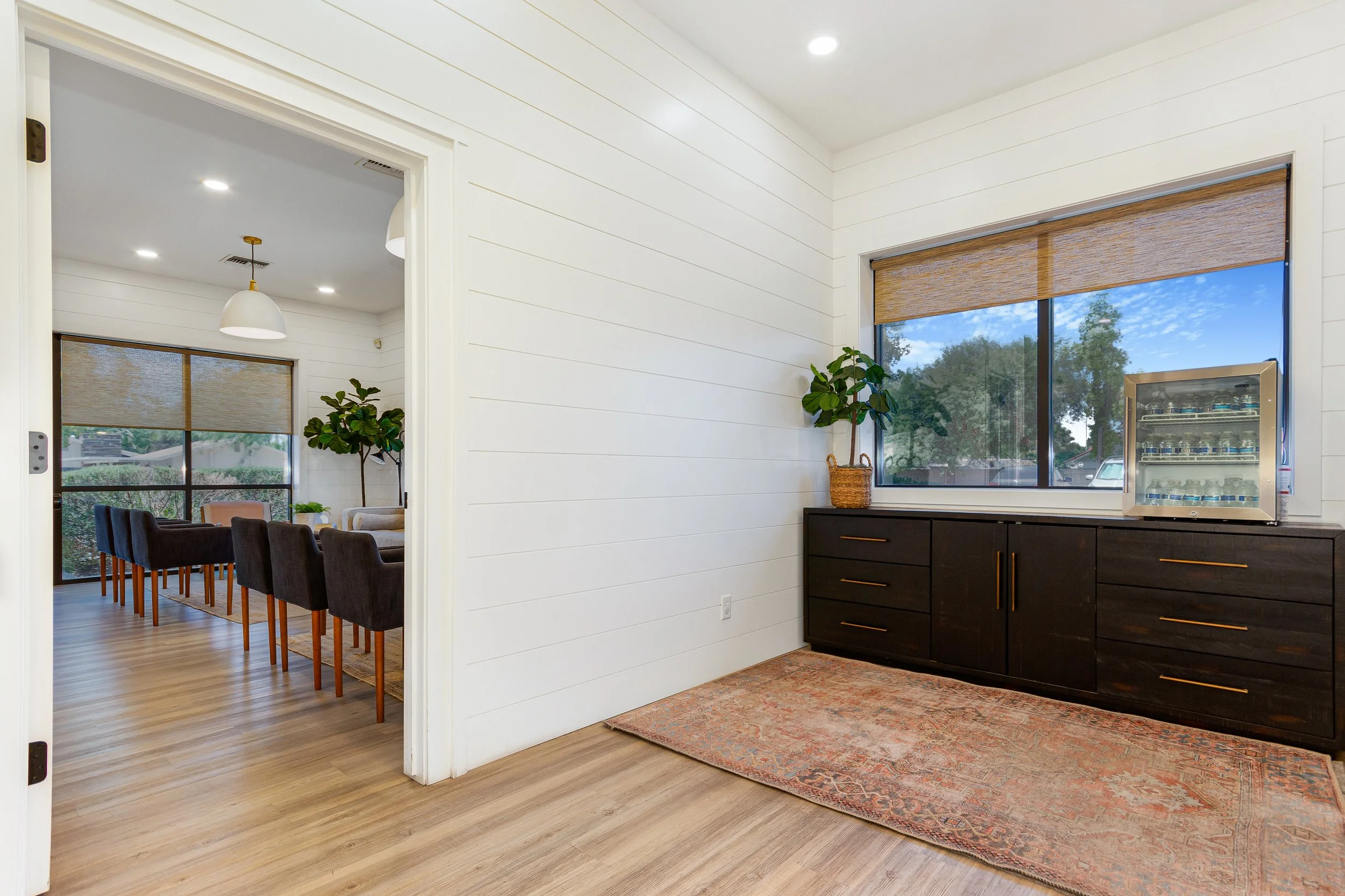 A cozy interior space with white shiplap walls, a large window with a brown roller shade, a black sideboard with gold handles, a potted plant in a woven basket, and a vending machine. Adjacent dining area visible through a doorway features a large wo