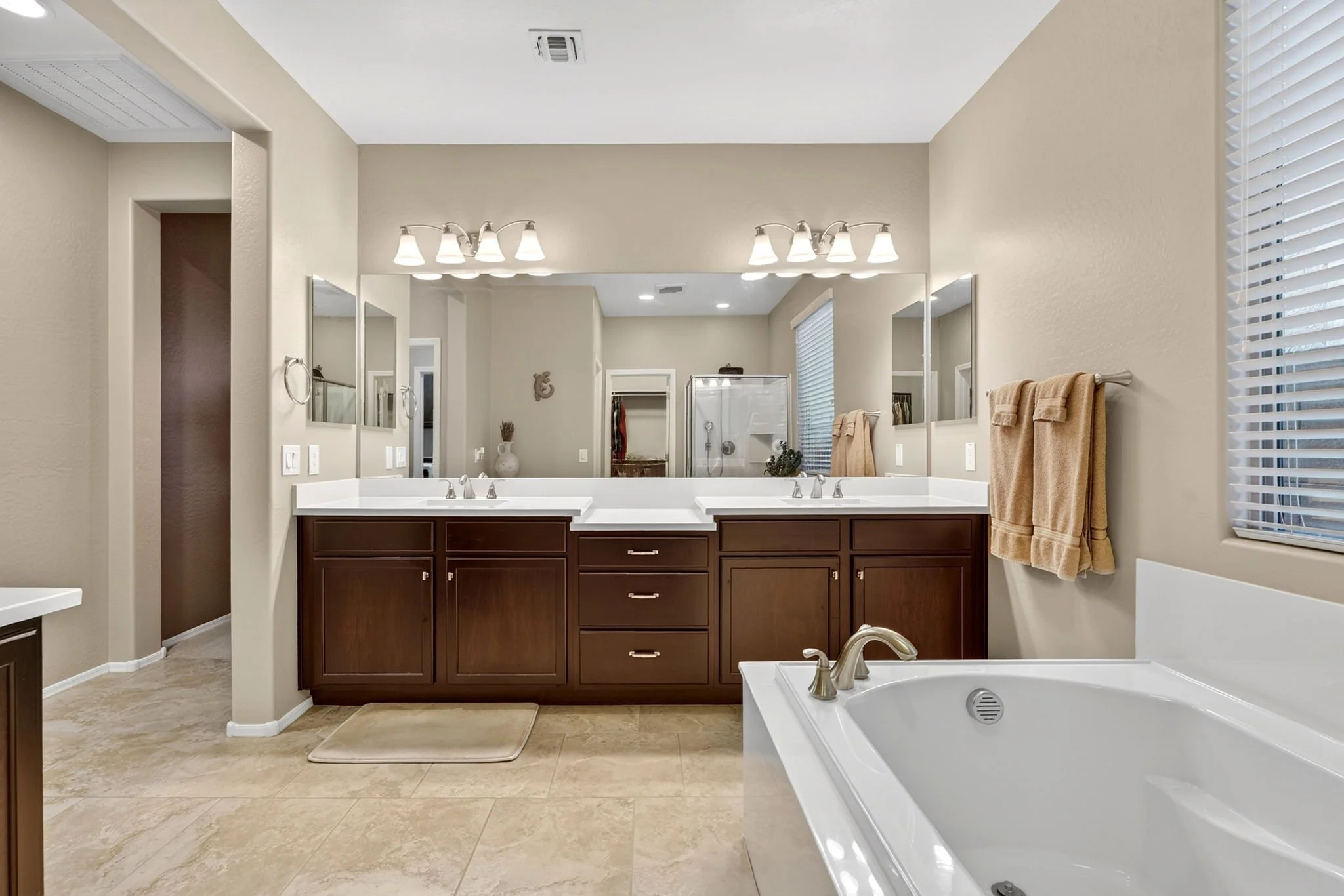 A spacious bathroom with beige walls, a double vanity with dark wood cabinets, a large mirror, beige towels, a bathtub, and a shower area with glass doors.